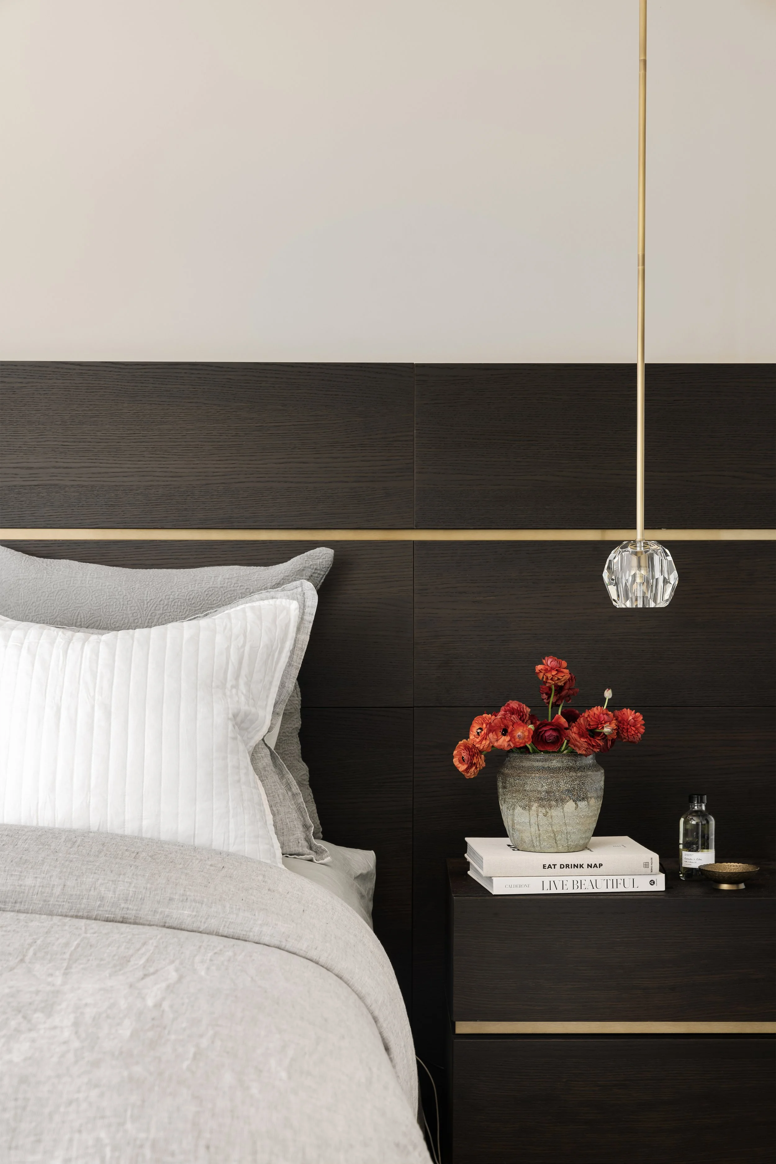 A bedroom scene showing part of a bed with white and gray pillows, a dark wood headboard with gold accents, a black nightstand with a vase of red flowers, books, a small bottle, and a dish, and a hanging pendant light with a crystal element.