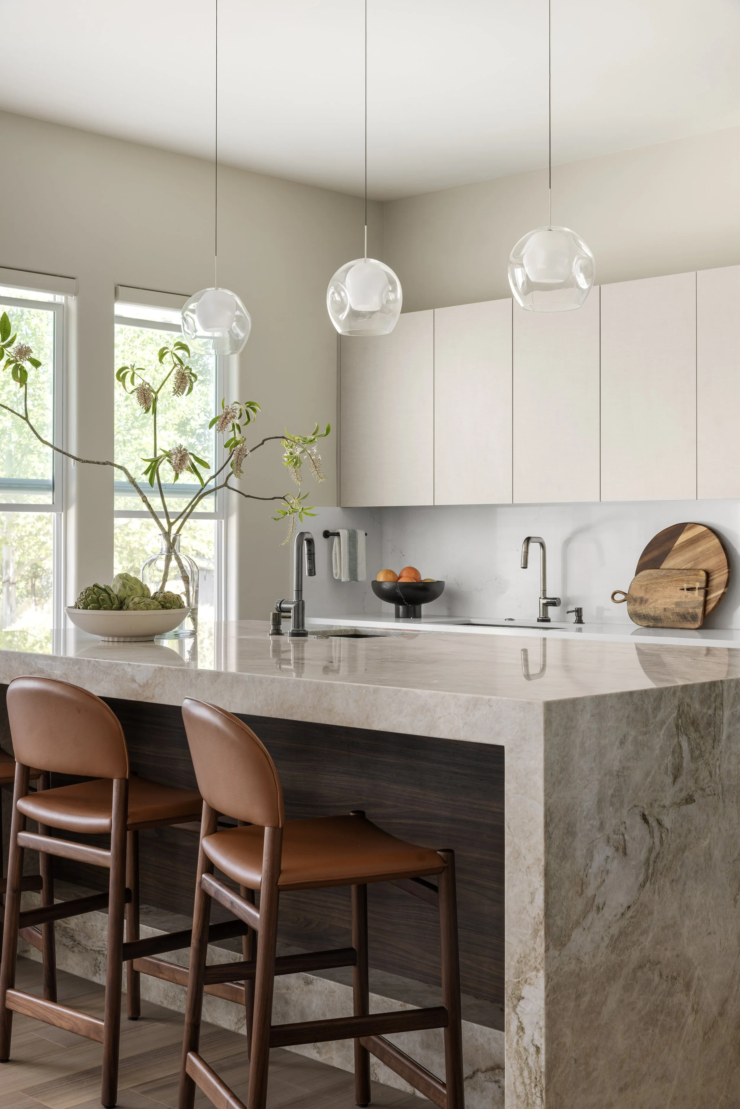 Modern kitchen with marble island, pendant lights, white cabinetry, and brown leather stools.