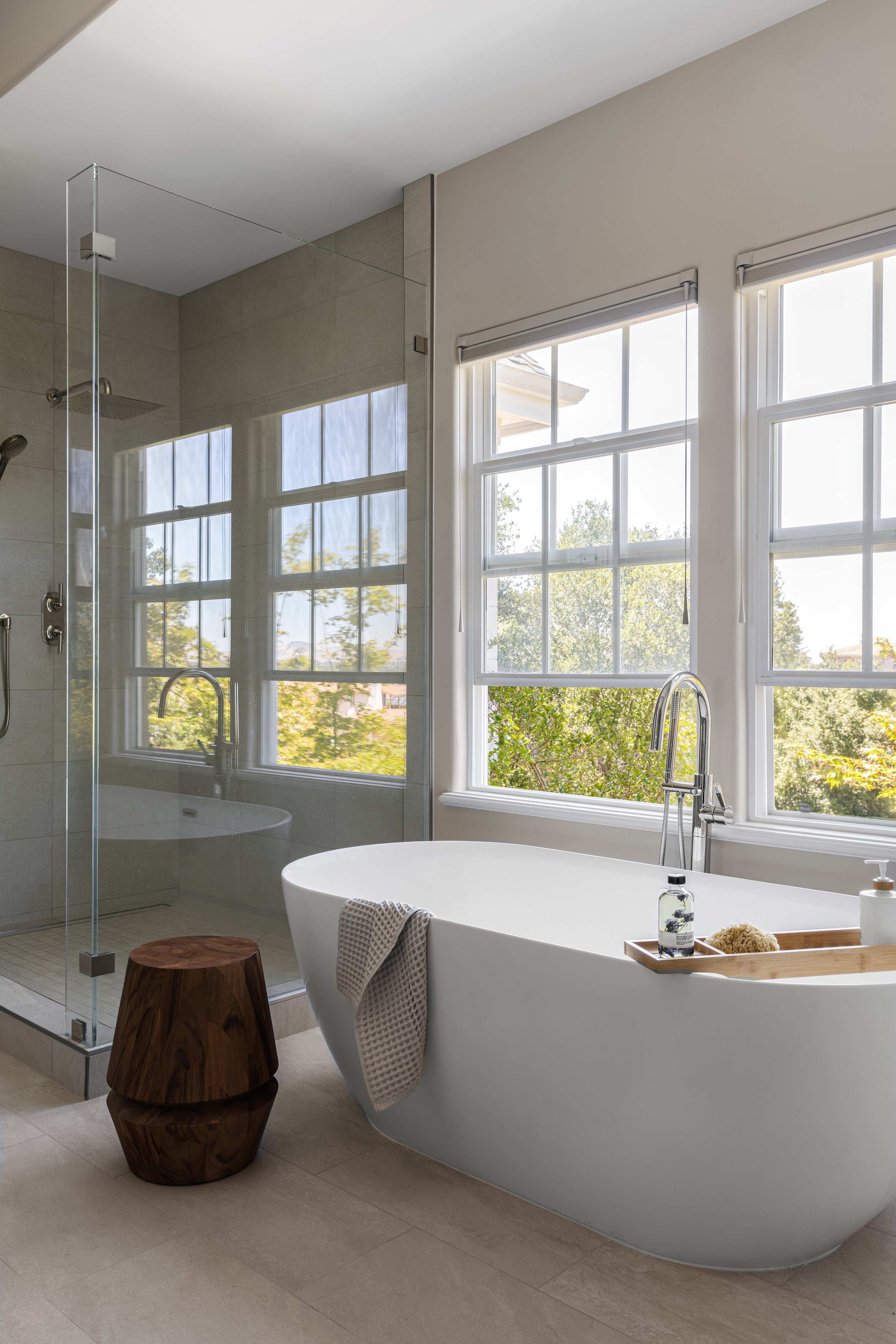 Modern bathroom with a white bathtub near large windows showing greenery outside, a wooden stool, and a glass-enclosed shower area.