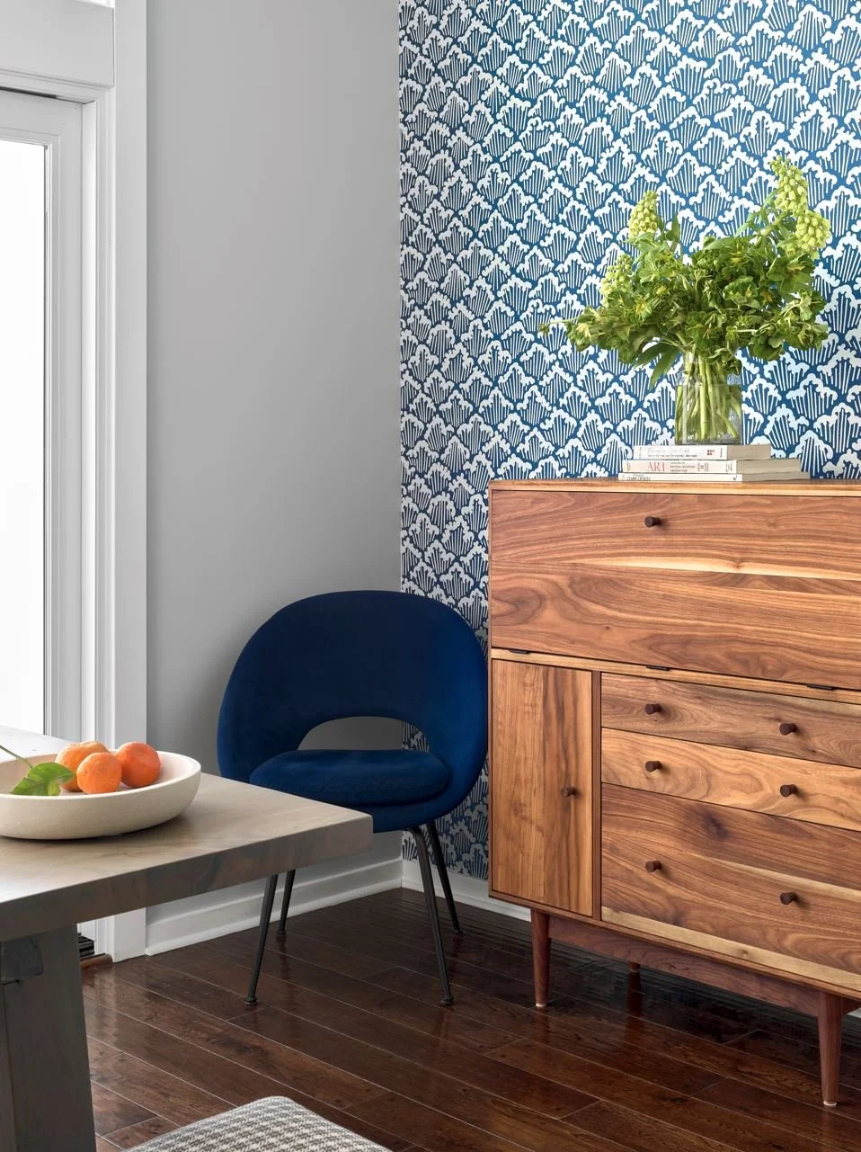 Interior of a room with a blue patterned wallpaper, a wooden dresser with a plant and books on top, a dark blue chair, a white table with a bowl of fruit, and hardwood floors.