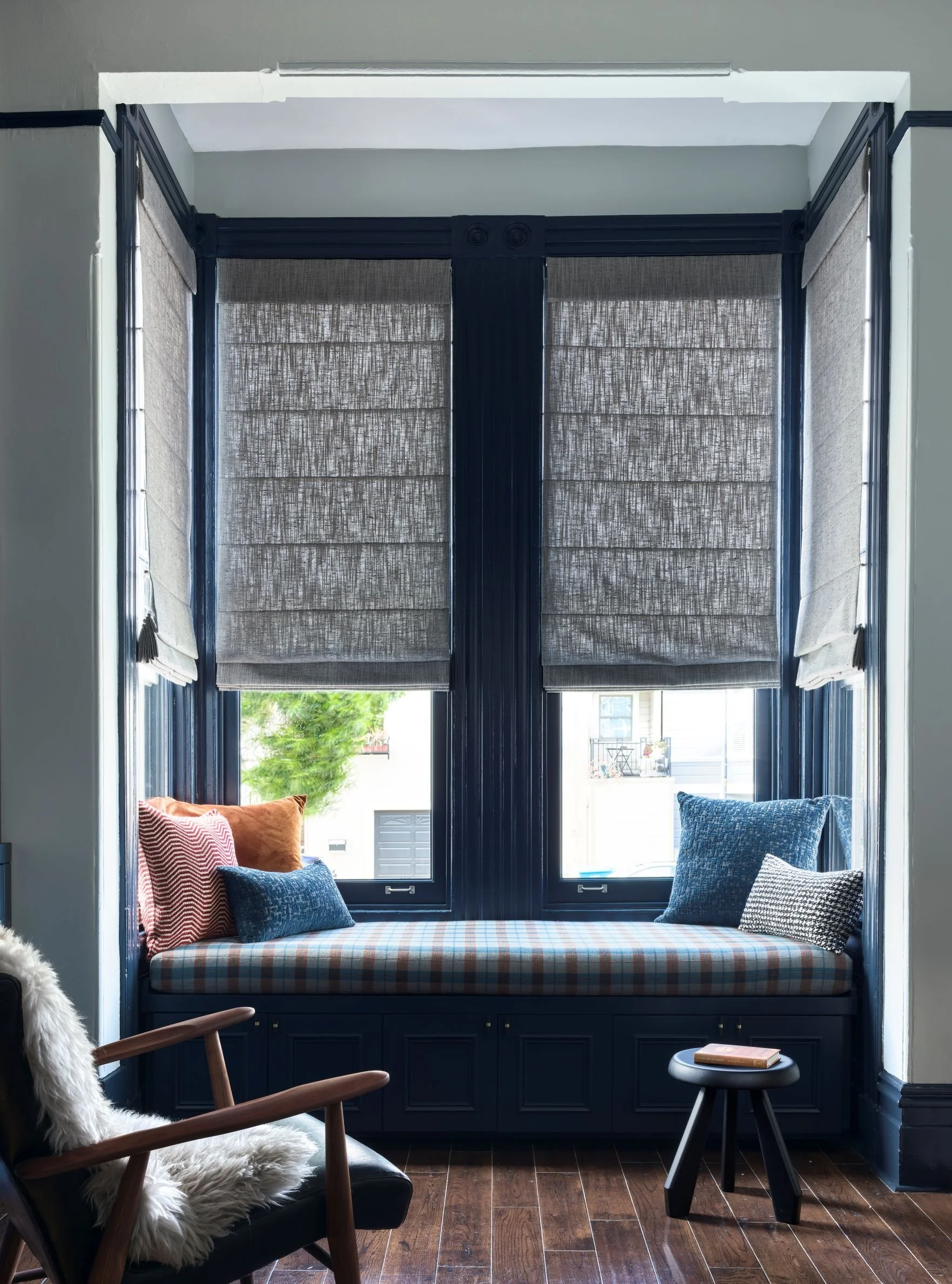 A window nook with a cushioned window seat, surrounded by blue window trim and gray Roman shades. Cushions and pillows are arranged on the seat, with a small black side table and a chair with a white furry throw nearby. The window overlooks a leafy s