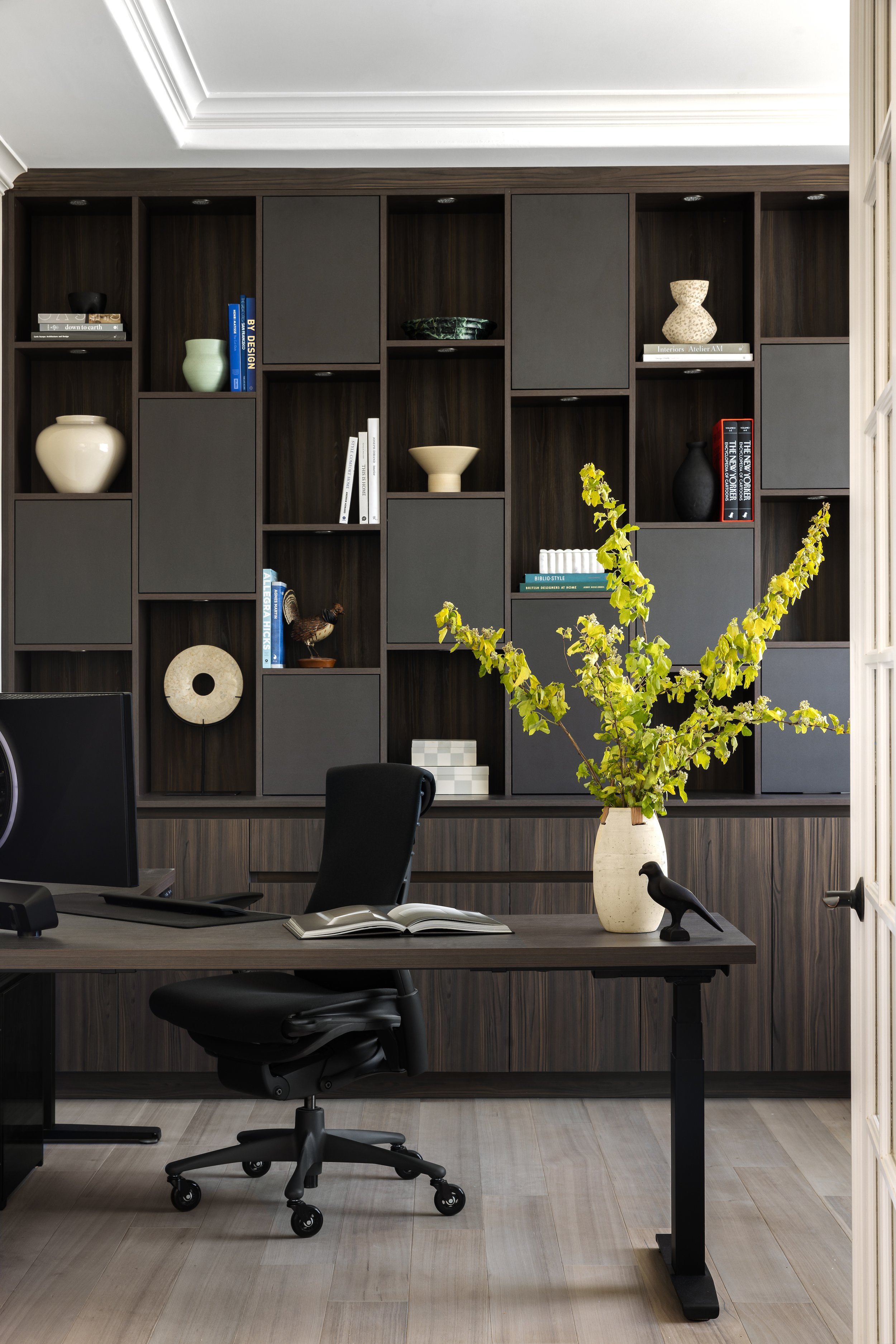 Modern office with dark wooden desk, black ergonomic chair, and decorative shelving filled with vases, books, and art. A large plant in a cream vase and a black bird figurine are on the desk.