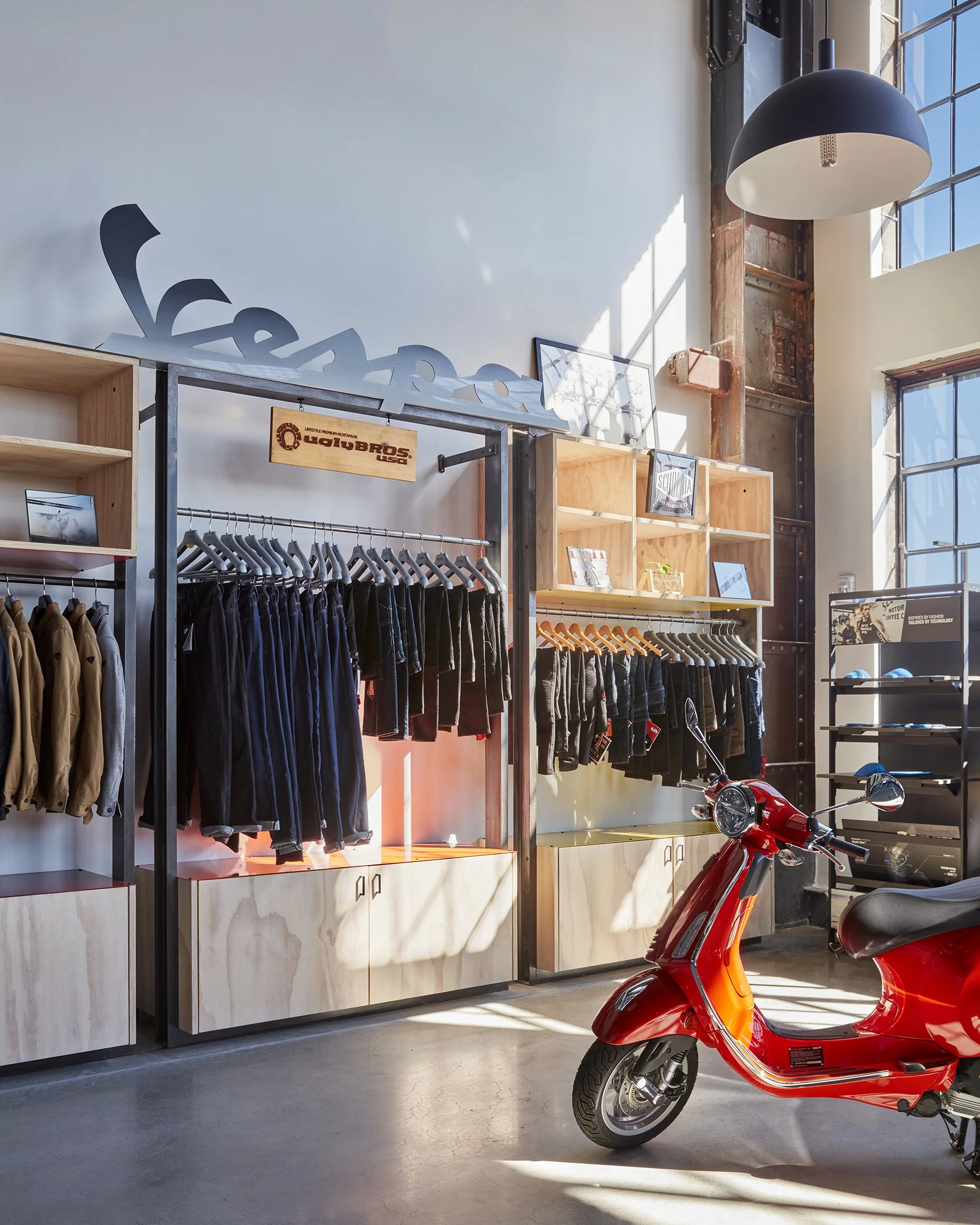 Interior of a retail store featuring clothing racks with jackets and pants, shelves with books and items, a red scooter on the floor, and large windows allowing sunlight to illuminate the space.