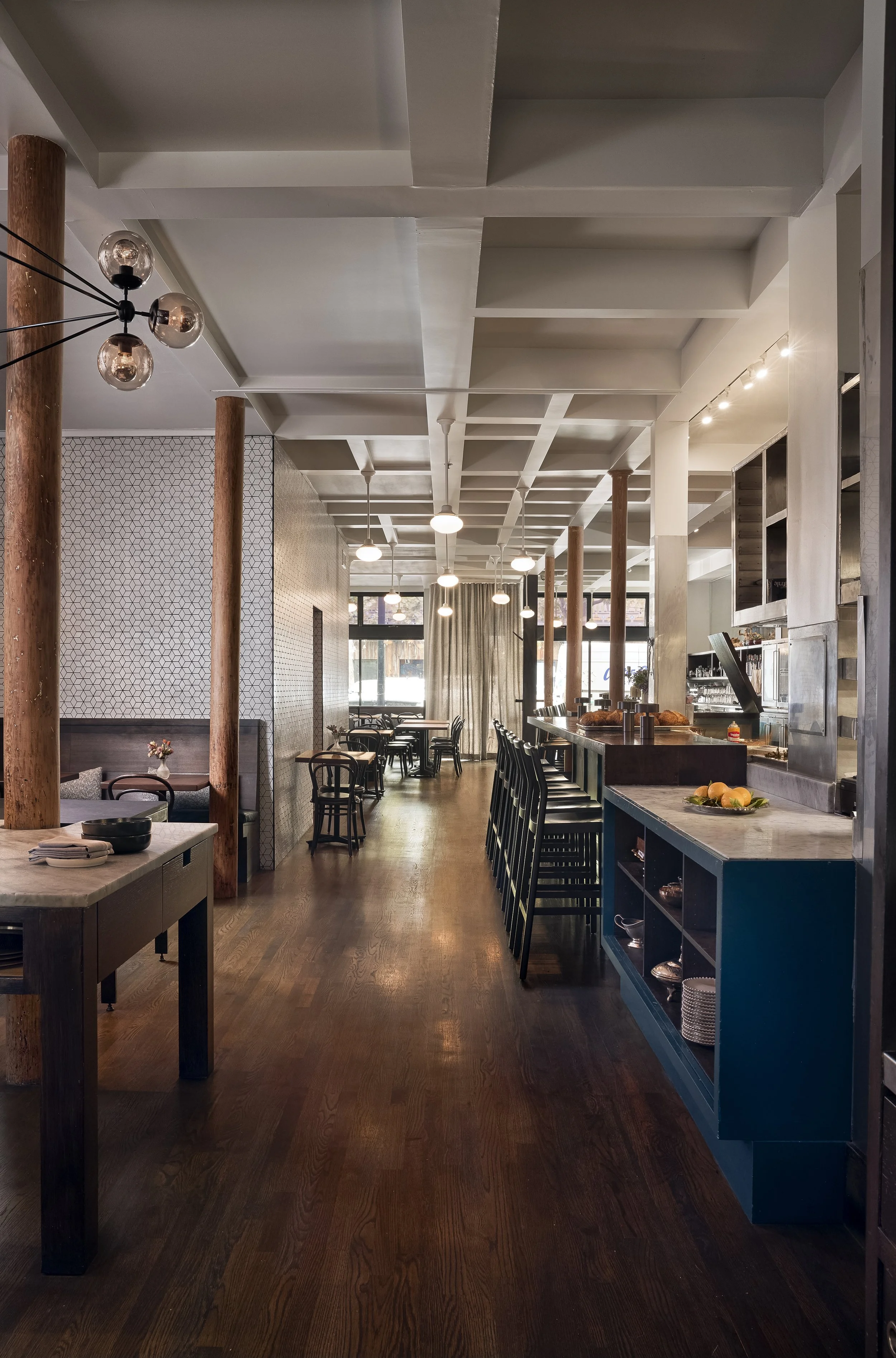 Interior of a modern restaurant or cafe with wooden floors, white ceiling with beams, pendant and chandelier lighting, a bar with tall chairs, and various tables and chairs along the windowed wall.