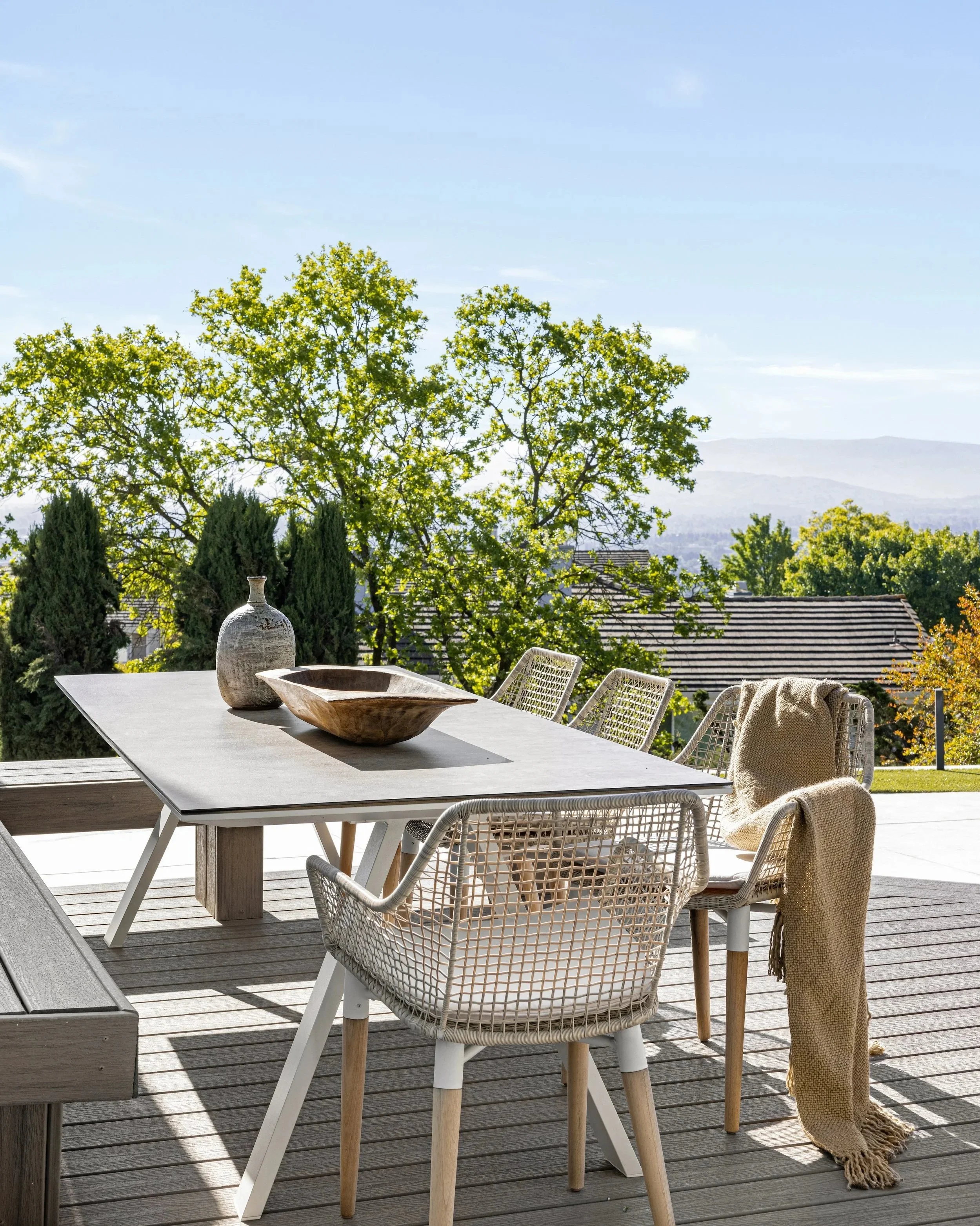 An outdoor patio with a dining table and chairs, set against a backdrop of trees and distant hills on a sunny day.