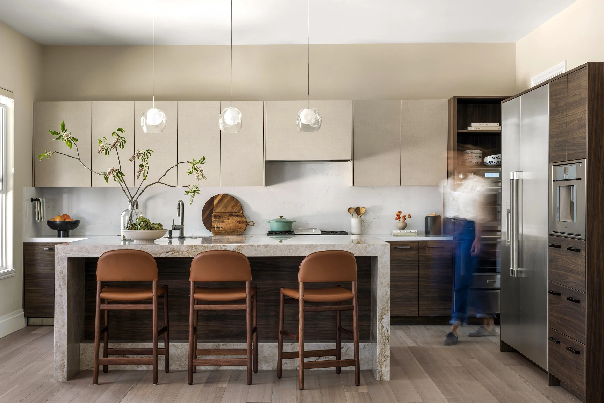 Modern kitchen with beige and wooden cabinets, marble countertops, three brown barstools, large vase with branches, and various kitchen accessories. Slight motion blur of a person walking.