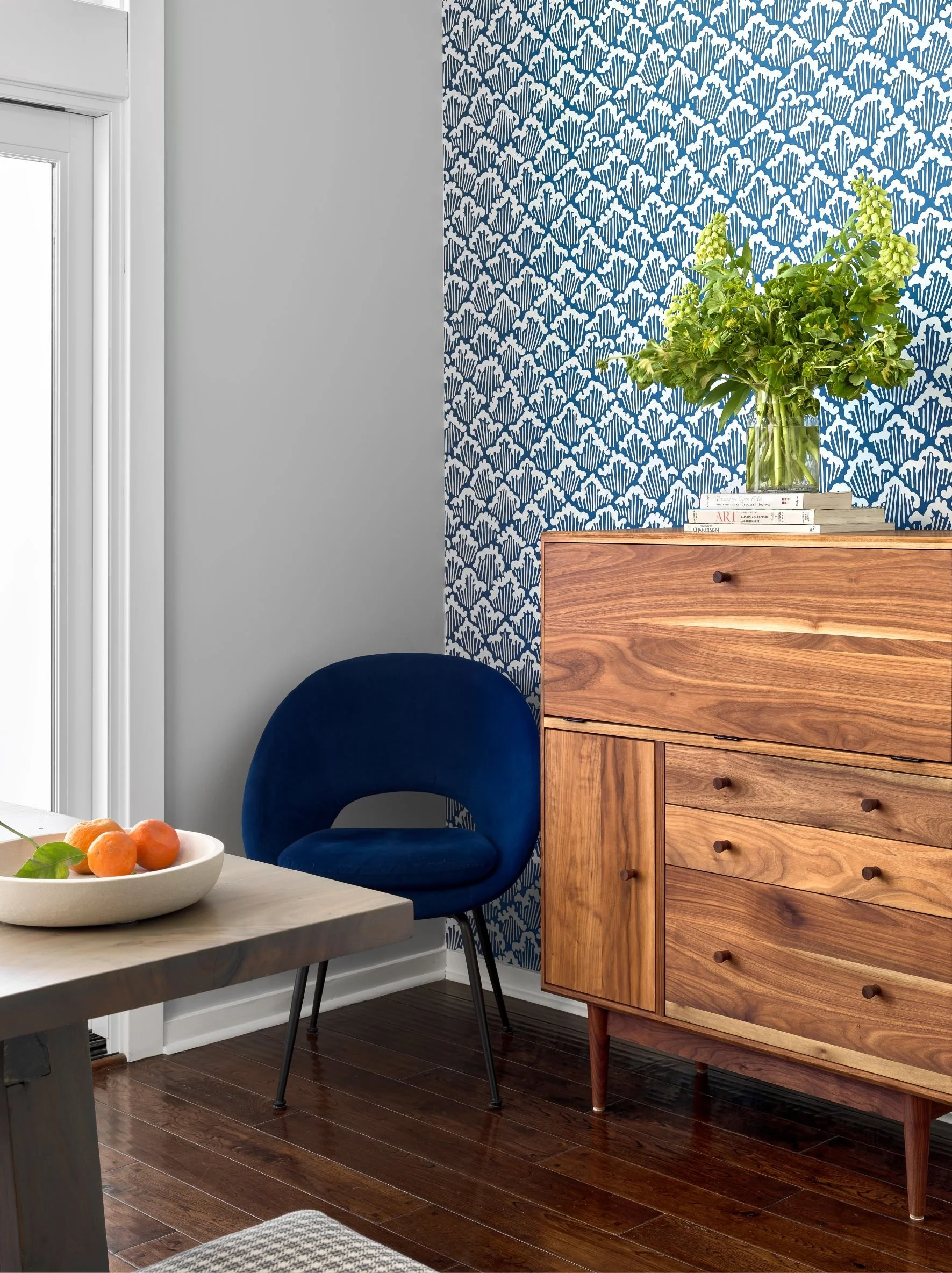 Interior of a room with a window, a wooden sideboard with a green plant and books, a blue velvet chair, a wooden table with a bowl of oranges, and patterned blue and white wallpaper.