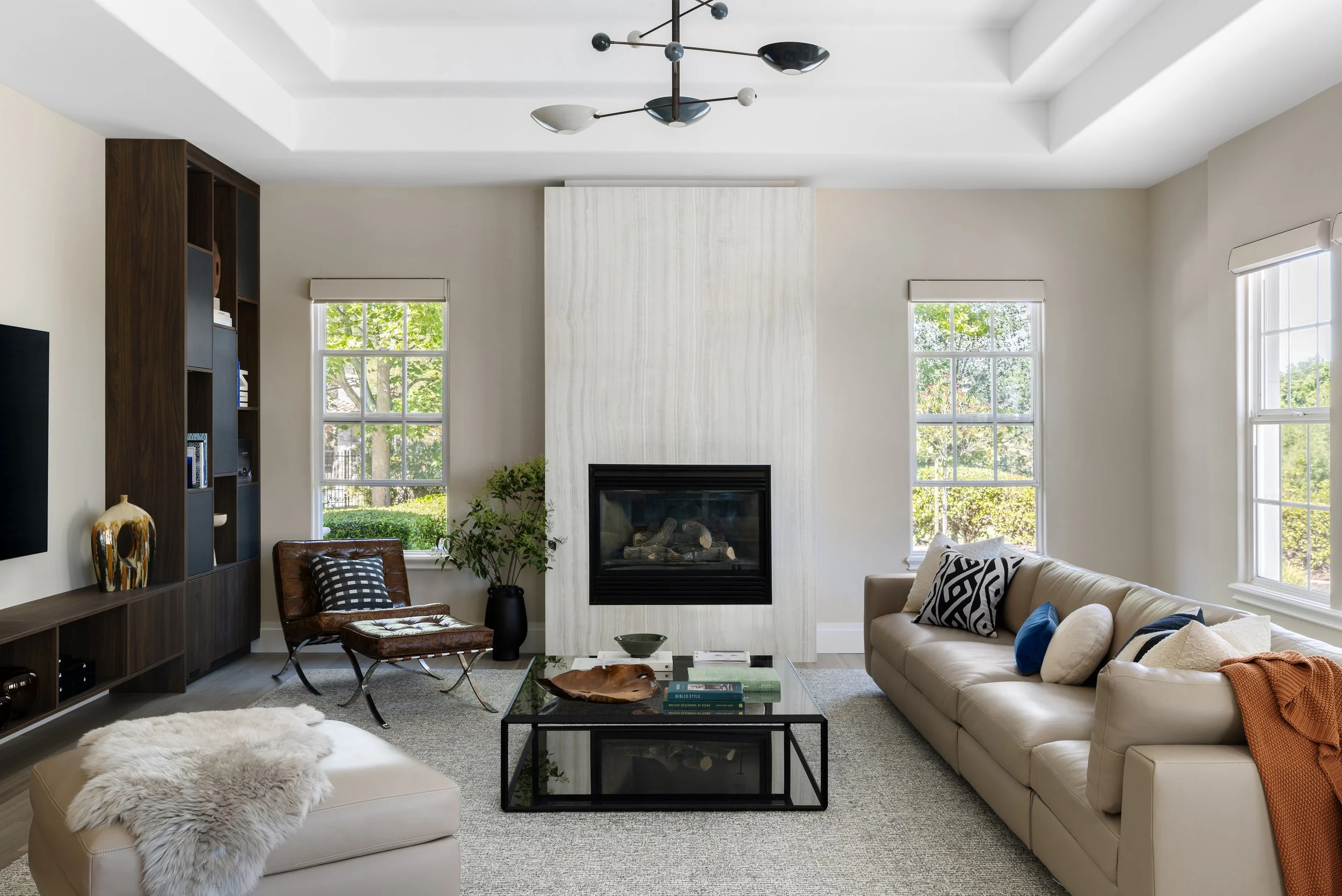 A modern living room with a white stone fireplace, large windows with views of greenery, a beige leather sofa with black, white, and blue pillows, a glass coffee table with books and decorative items, a brown leather chair with a black and white pill