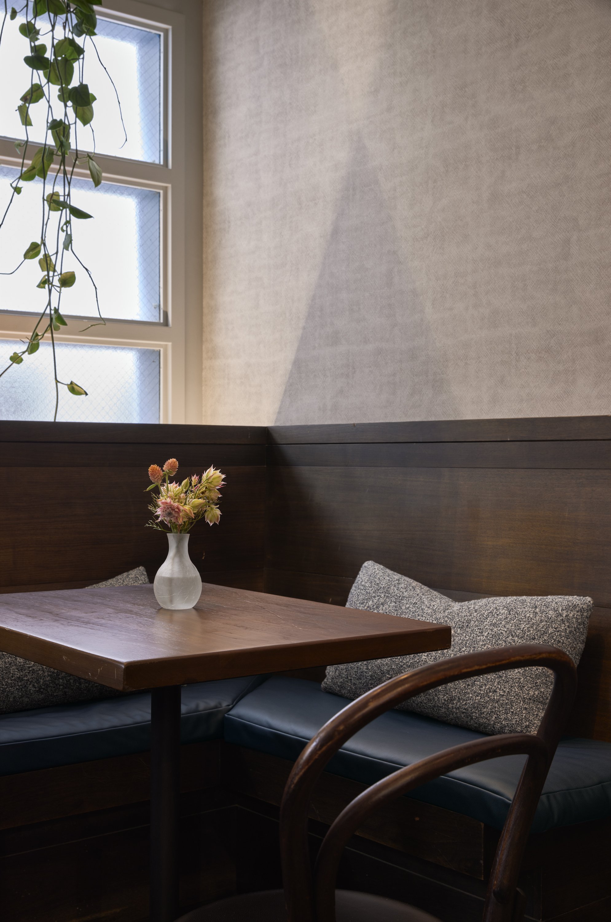 Corner of a cozy café or dining area with a wooden table, a small vase of pink and white flowers, grey cushions, and a dark wood bench. A large window with frosted glass allows natural light in.