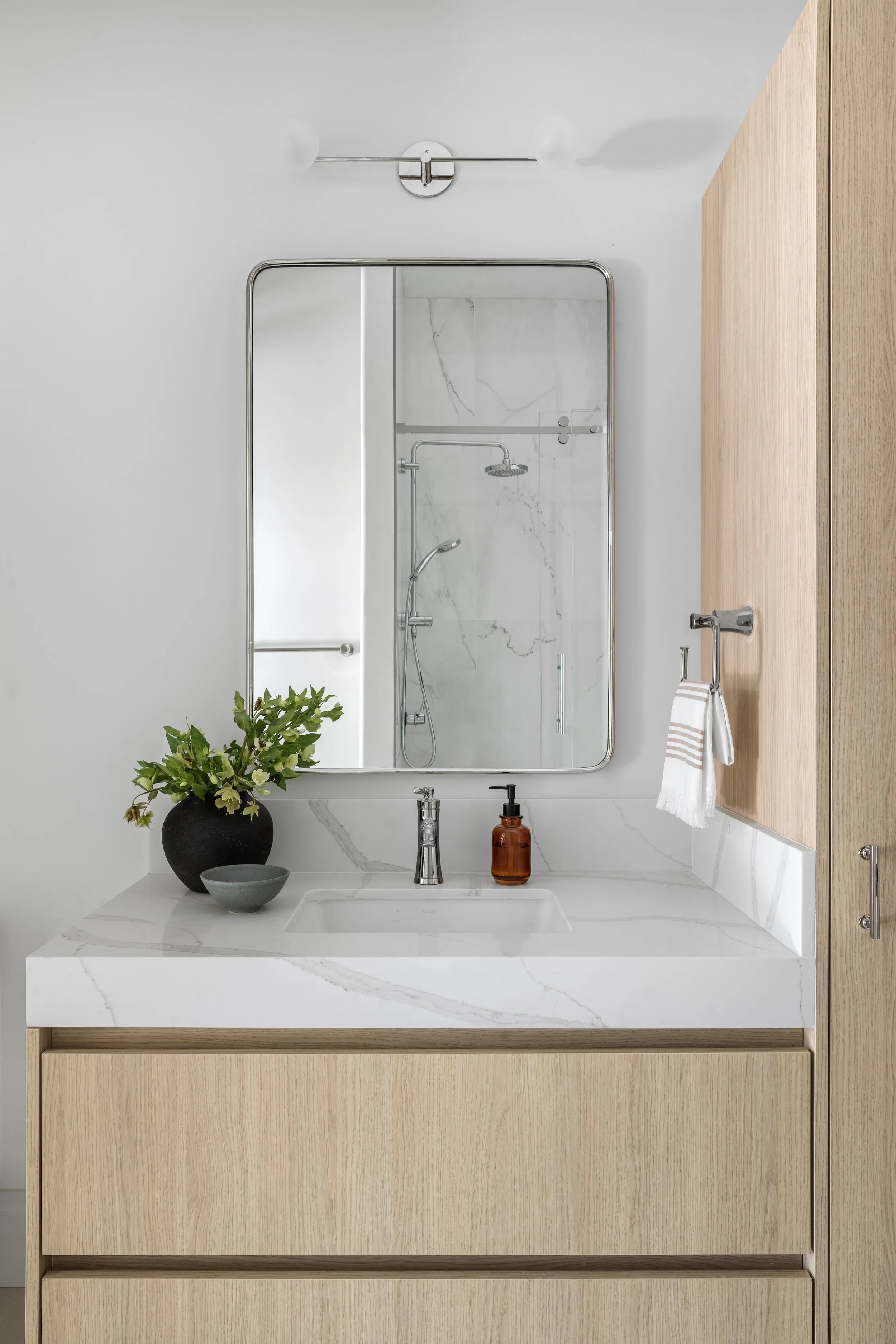 Modern bathroom vanity with a white marble countertop, a sink, a bronze soap dispenser, a black vase with green plants, a small bowl, a wall-mounted mirror, a wall-mounted light fixture, a wooden cabinet, and a towel hook with a towel. The bathroom h