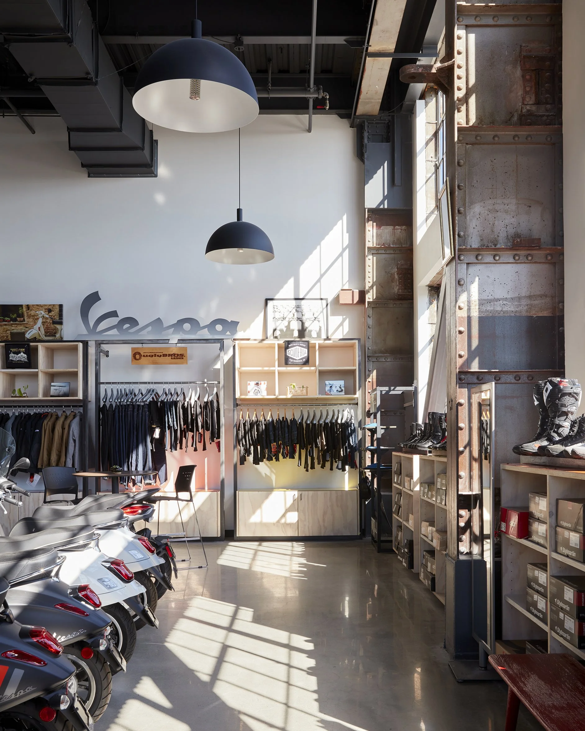Interior of a retail store showing motorcycles parked on the left, clothing racks with jackets and shirts, shelves with shoes and boxes, and large industrial-style metal wall on the right, with sunlight streaming through large windows.