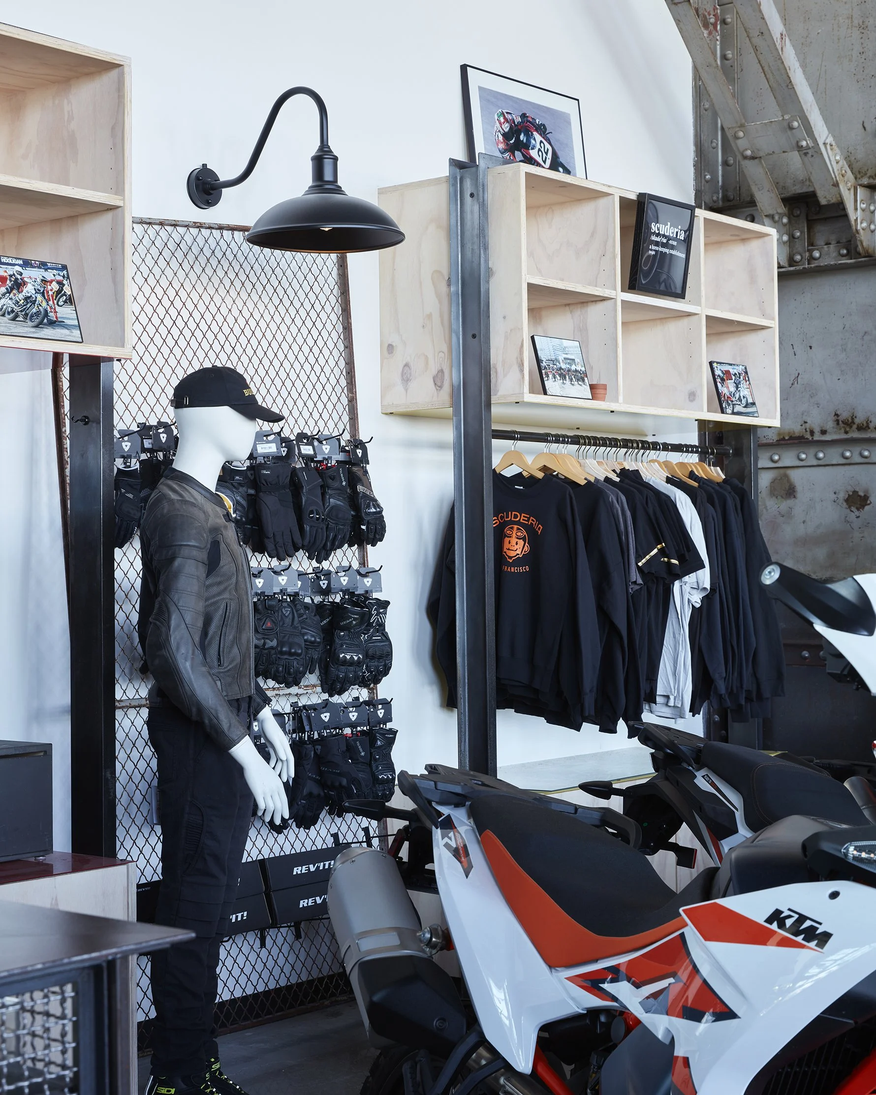 Motorcycle shop display with riding gear, a mannequin dressed in leather jacket and pants, gloves, helmets, black and orange KTM motorcycle, and framed motorcycle photos.