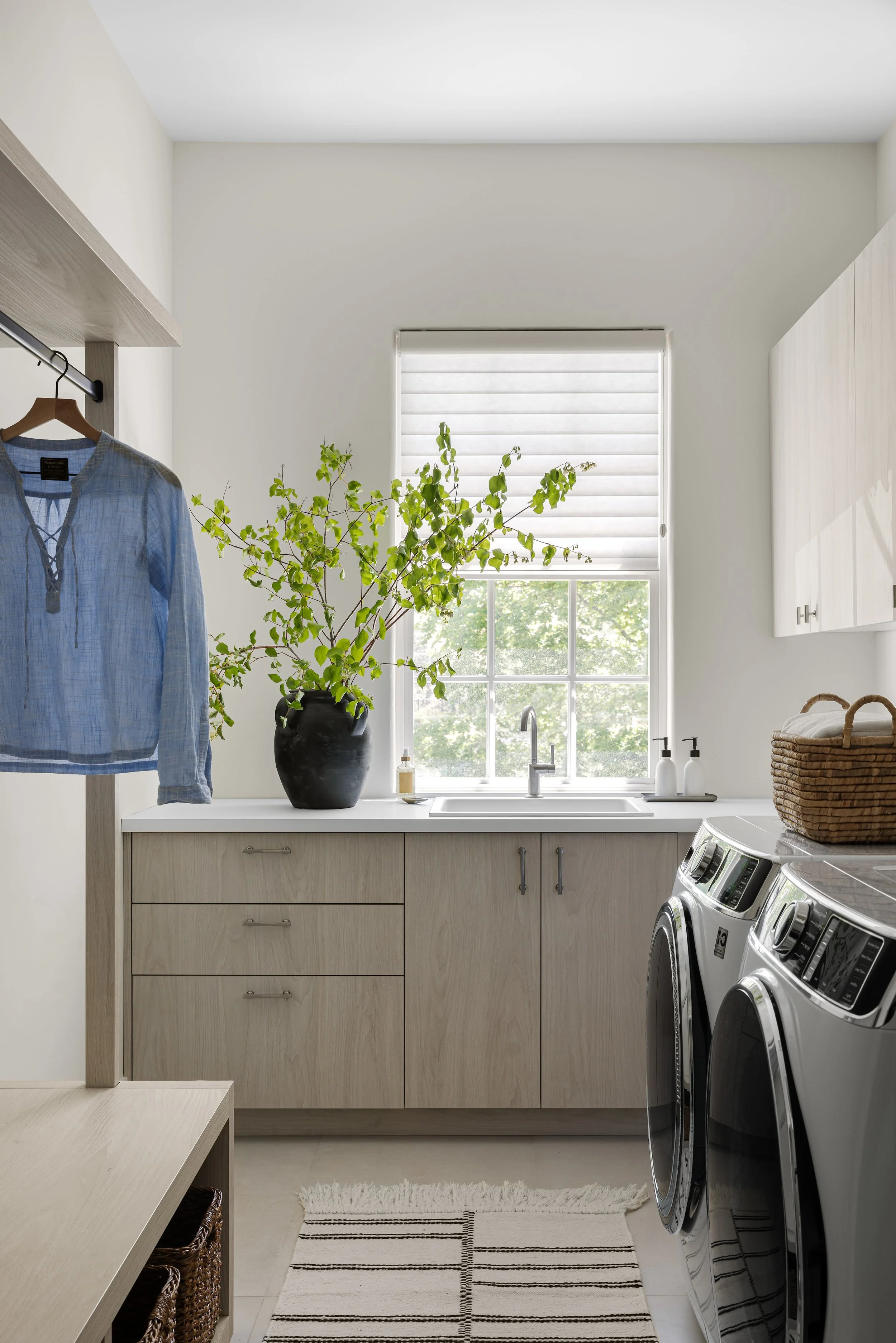 Laundry room with washer and dryer, a window with blinds, a large black vase with green branches, and white cabinets.