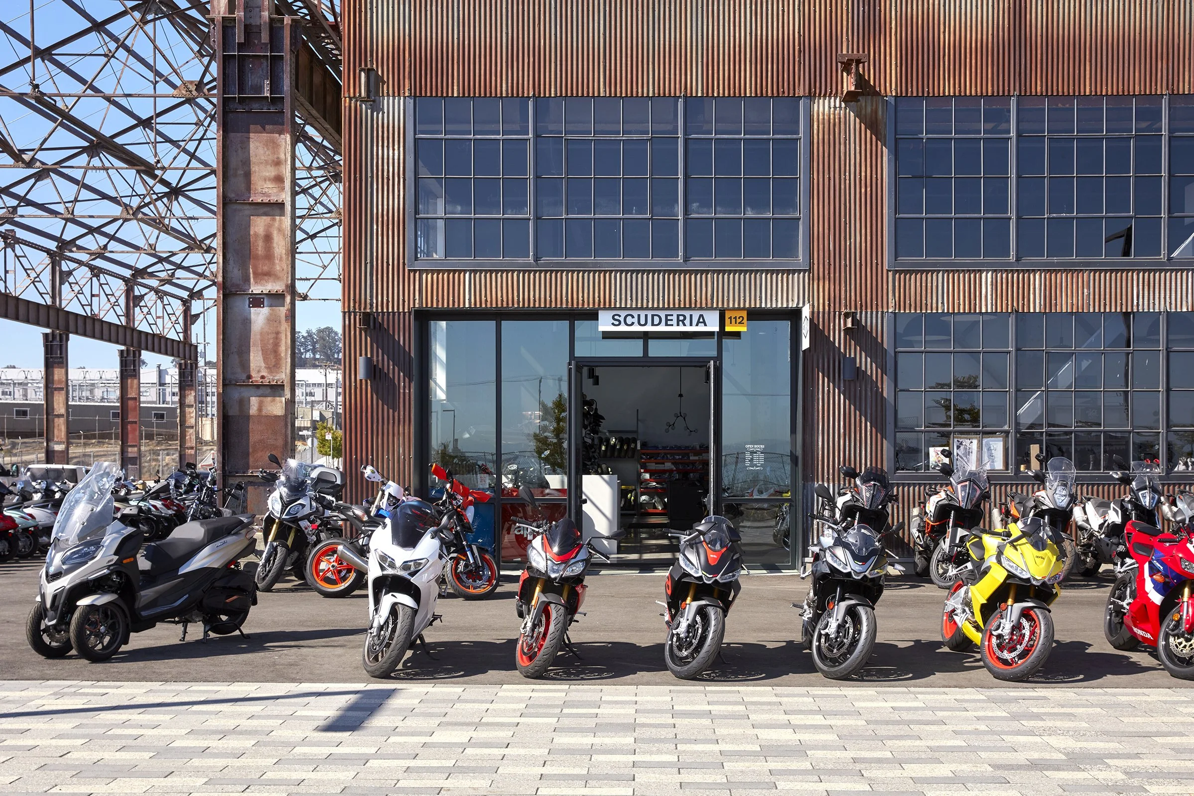 A motorcycle dealership with a variety of motorcycles parked outside, in front of a rusty metal building with large glass windows and a sign that reads "SCUDERIA".
