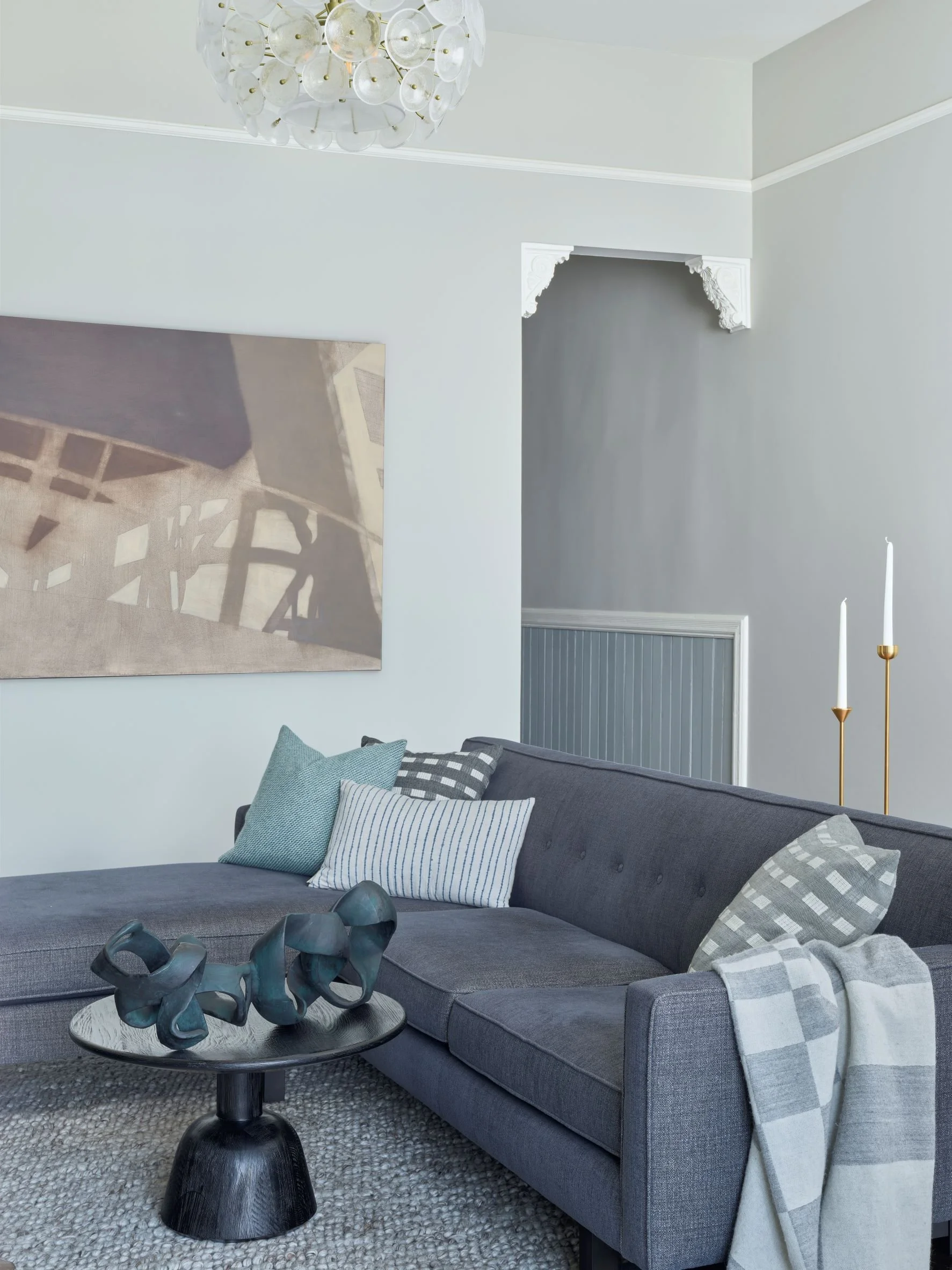 A living room with a gray sectional sofa decorated with various cushions, a black side table with a decorative sculpture, a textured rug, and a white wall with a large abstract painting. There is a gold candle holder with white candles and a modern c
