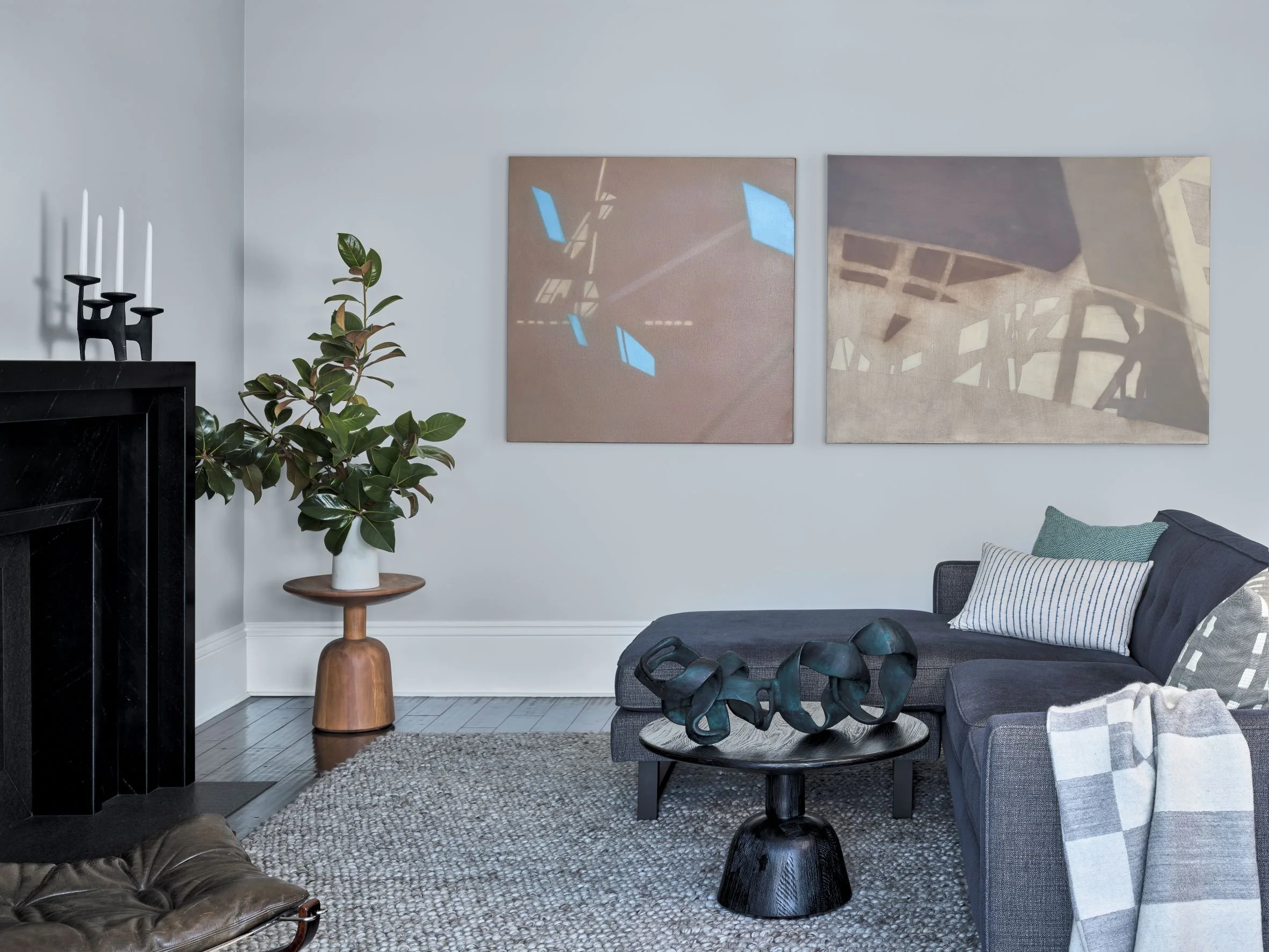 Living room with gray sectional sofa, abstract art on wall, black coffee table with sculpture, green plant in white vase, and fireplace with black mantel and white candles.