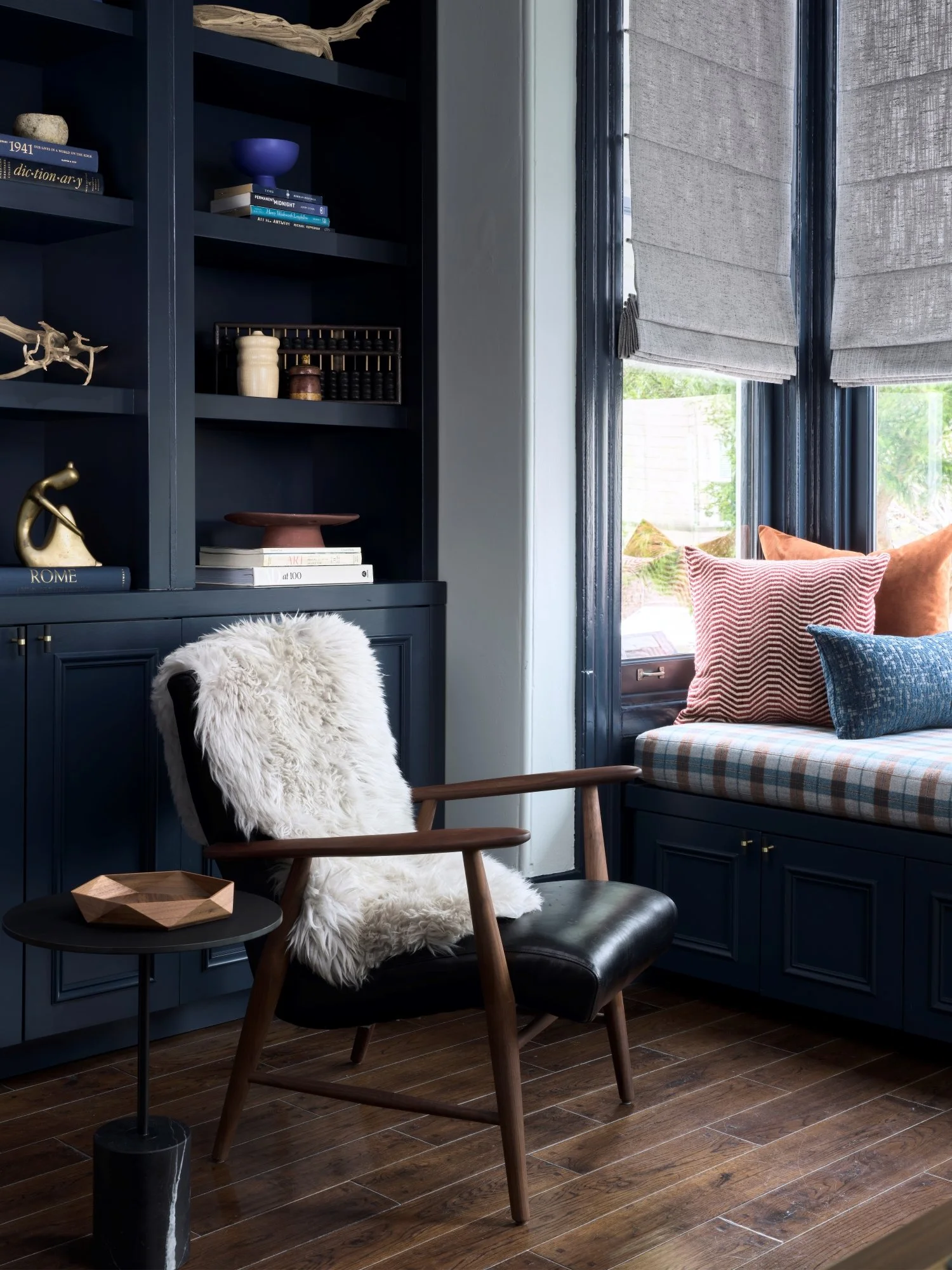 Cozy corner of a room with a dark blue bookcase, a black leather chair with a white faux fur throw, a small black side table with a wooden tray, and a window seat with colorful pillows and plaid cushion, natural light streaming in.