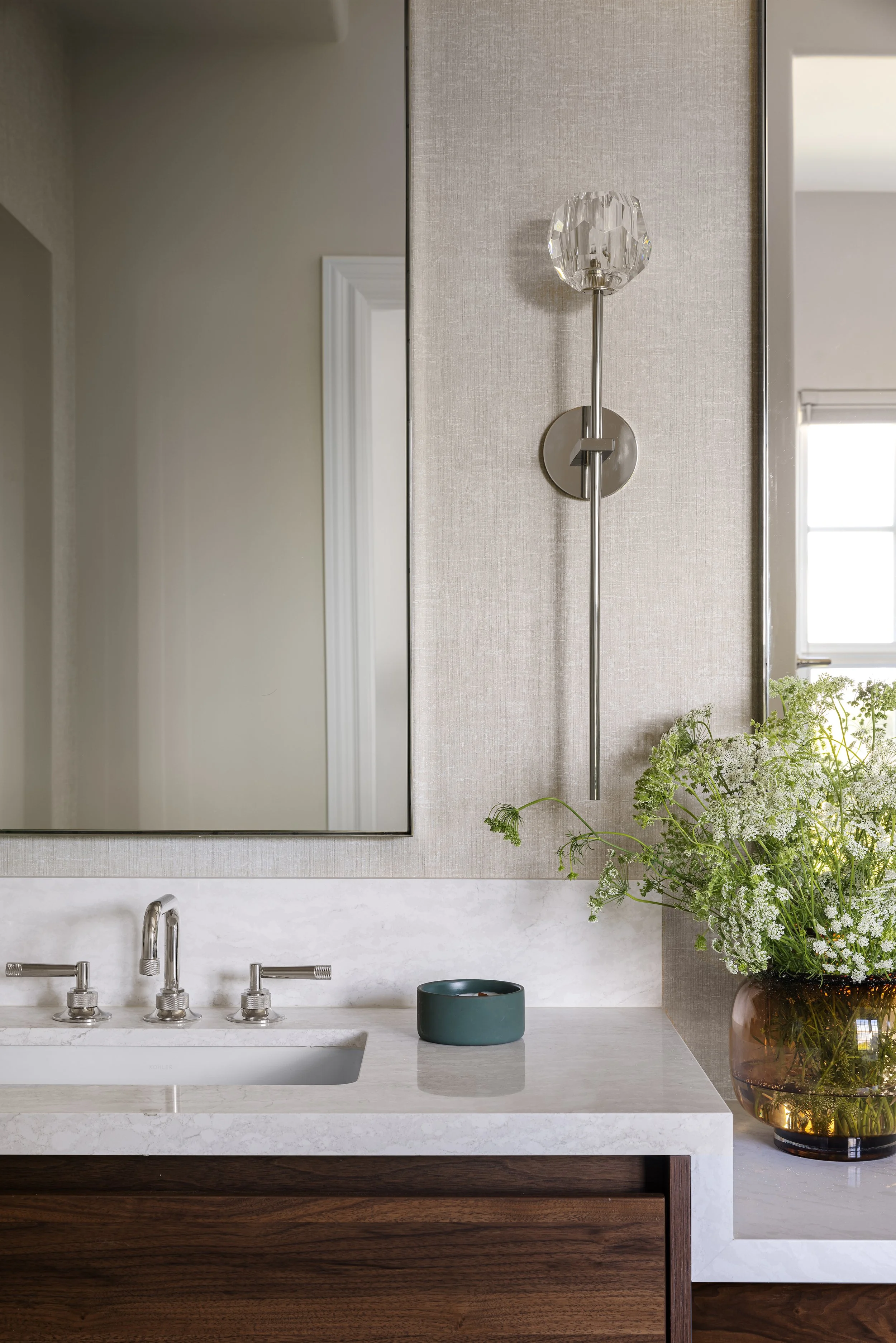 A modern bathroom with a white marble countertop, a small dark bowl, a large clear flower vase with white flowers, a large mirror, a textured beige wall, a contemporary wall sconce with a crystal globe, a window in the background, and a wooden cabine