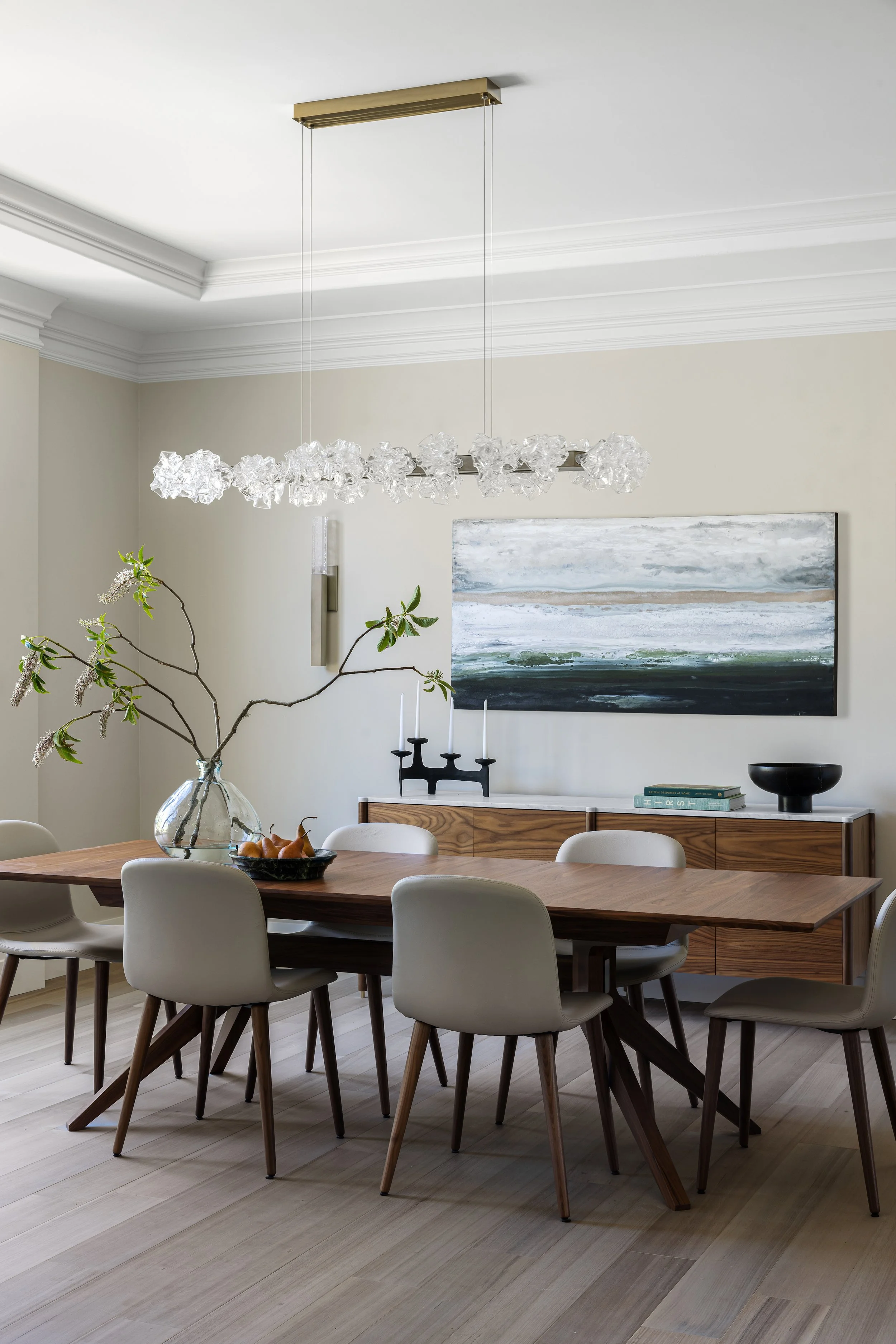 Modern dining room with a wooden table, six beige chairs, a glass vase with branches, a black bowl with fruit, a wooden sideboard, a large abstract painting, and a chandelier.