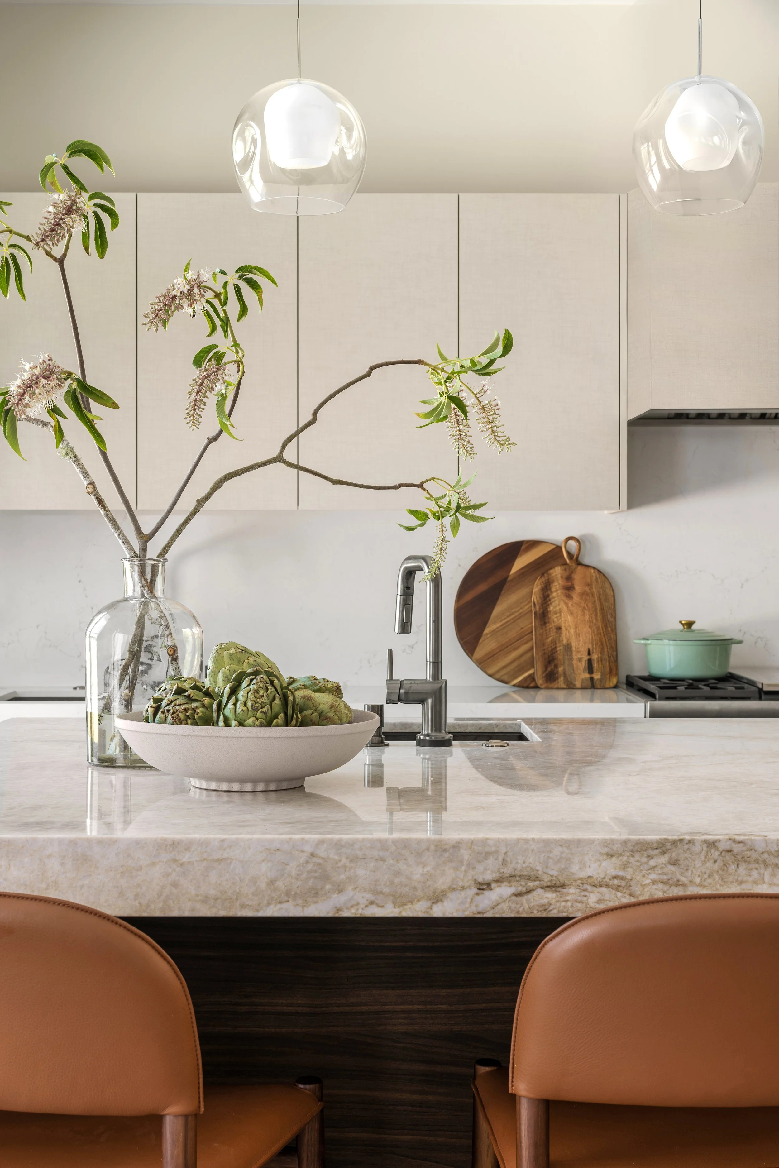 A modern kitchen with a marble island, a large glass vase with branches and leaves, artichokes in a bowl, and wooden cutting boards. Overhead are two glass pendant lights.