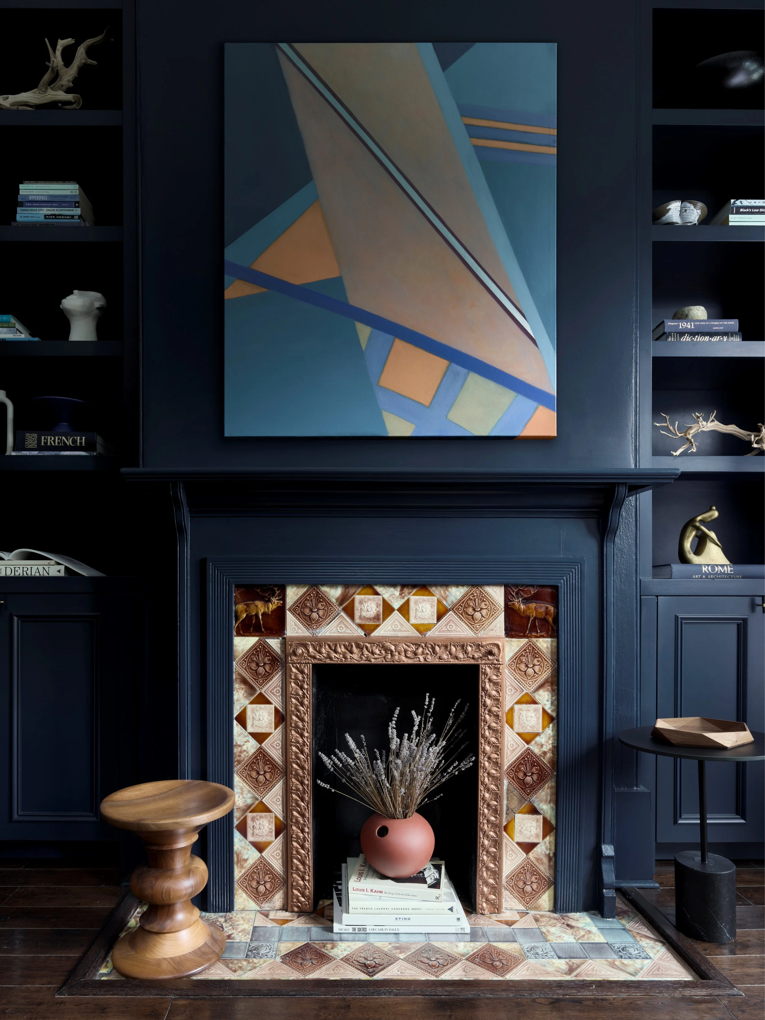 Interior of a room with a dark blue fireplace, decorated with patterned tiles, topped by a large abstract painting, with shelves holding books and decorative objects.