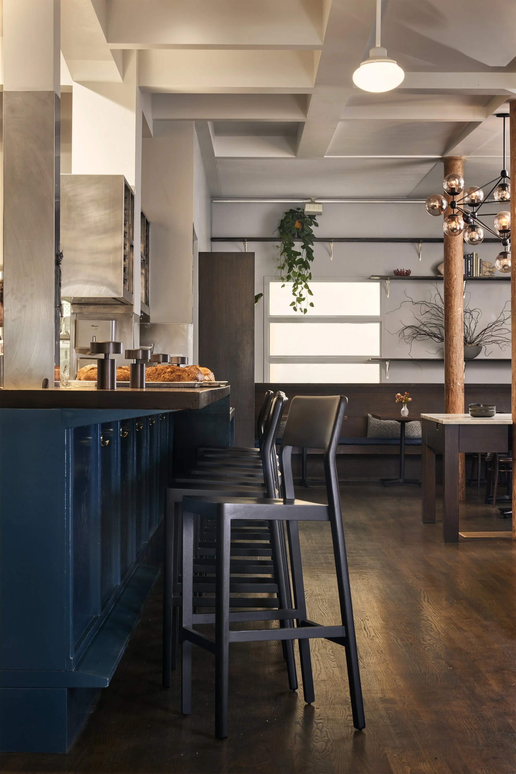 Interior view of a modern cafe with dark wood floors, black bar stools, and a teal-colored bar counter. There are two layers of shelves on the wall with decorative items, a small table with a flower vase, and hanging lighting fixtures. Natural light 
