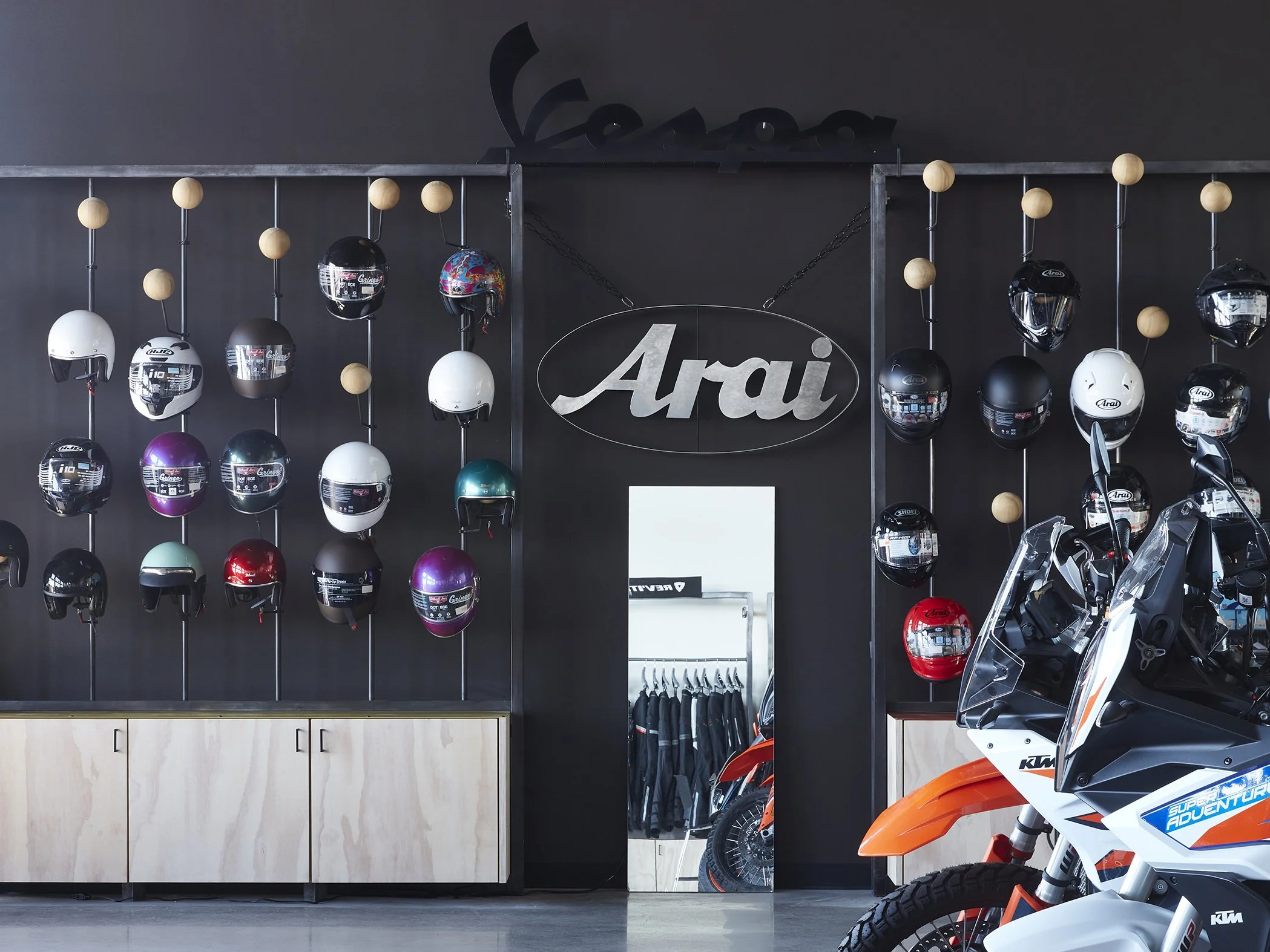 Display of motorcycle helmets on a wall with a large Arai logo above and an orange and white KTM dirt bike in the foreground inside a motorcycle gear shop.