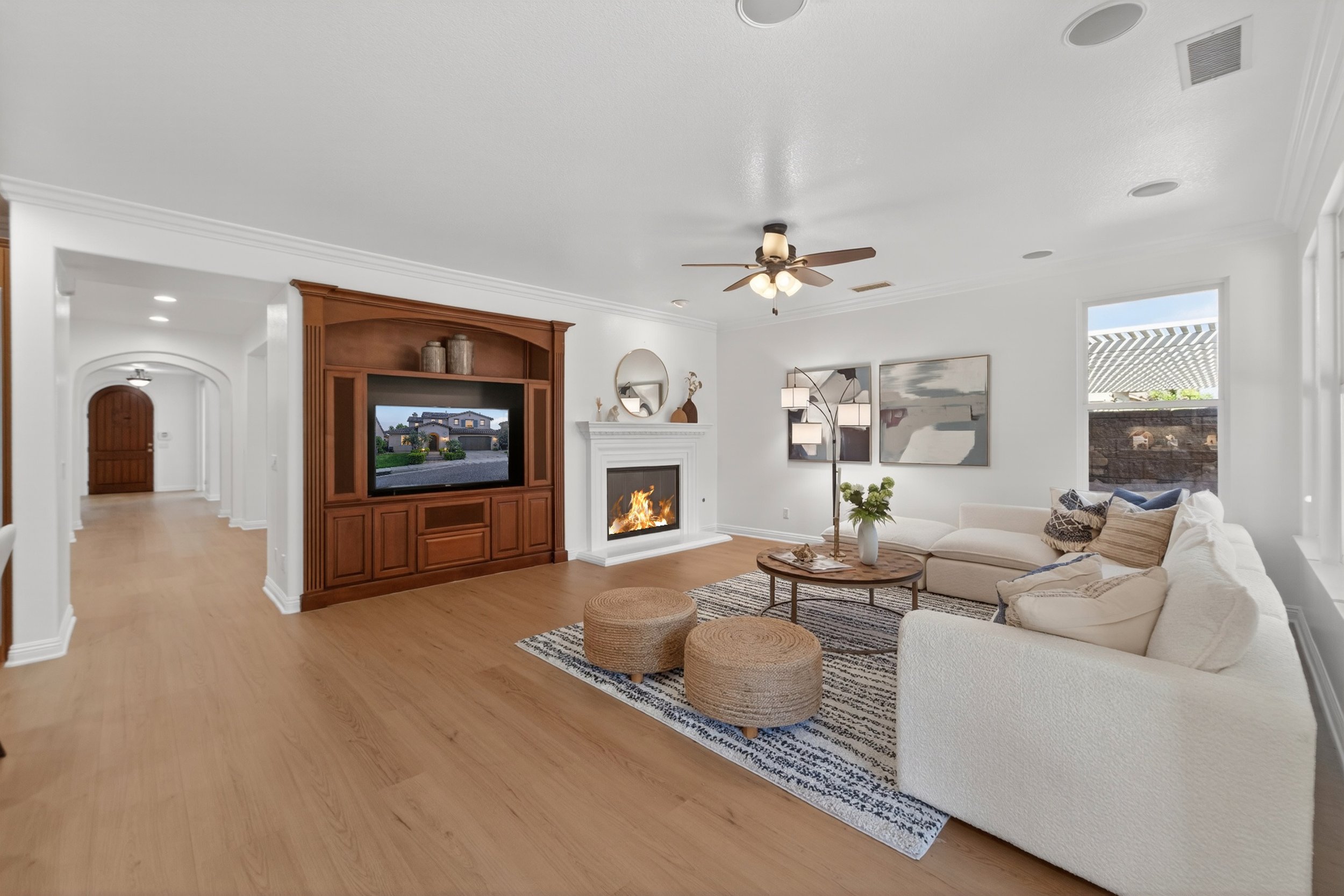 A living room with white walls, a fireplace, a ceiling fan, and a flat-screen TV surrounded by a wooden entertainment center. There are white sofas, a coffee table, and wall art, with large windows letting in natural light.
