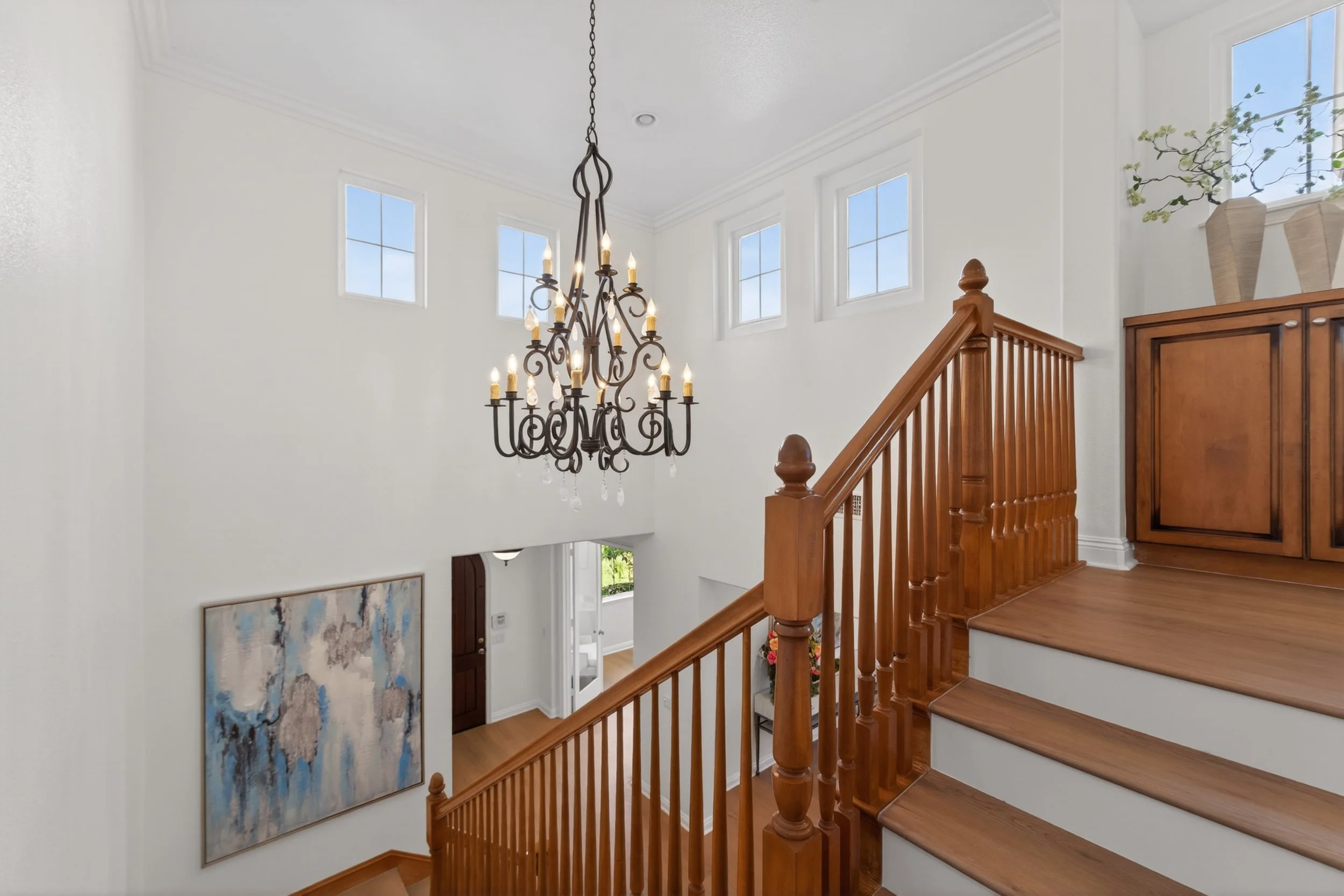 Interior view of a stairway landing with a chandelier, high windows, potted plants, and a wooden cabinet.