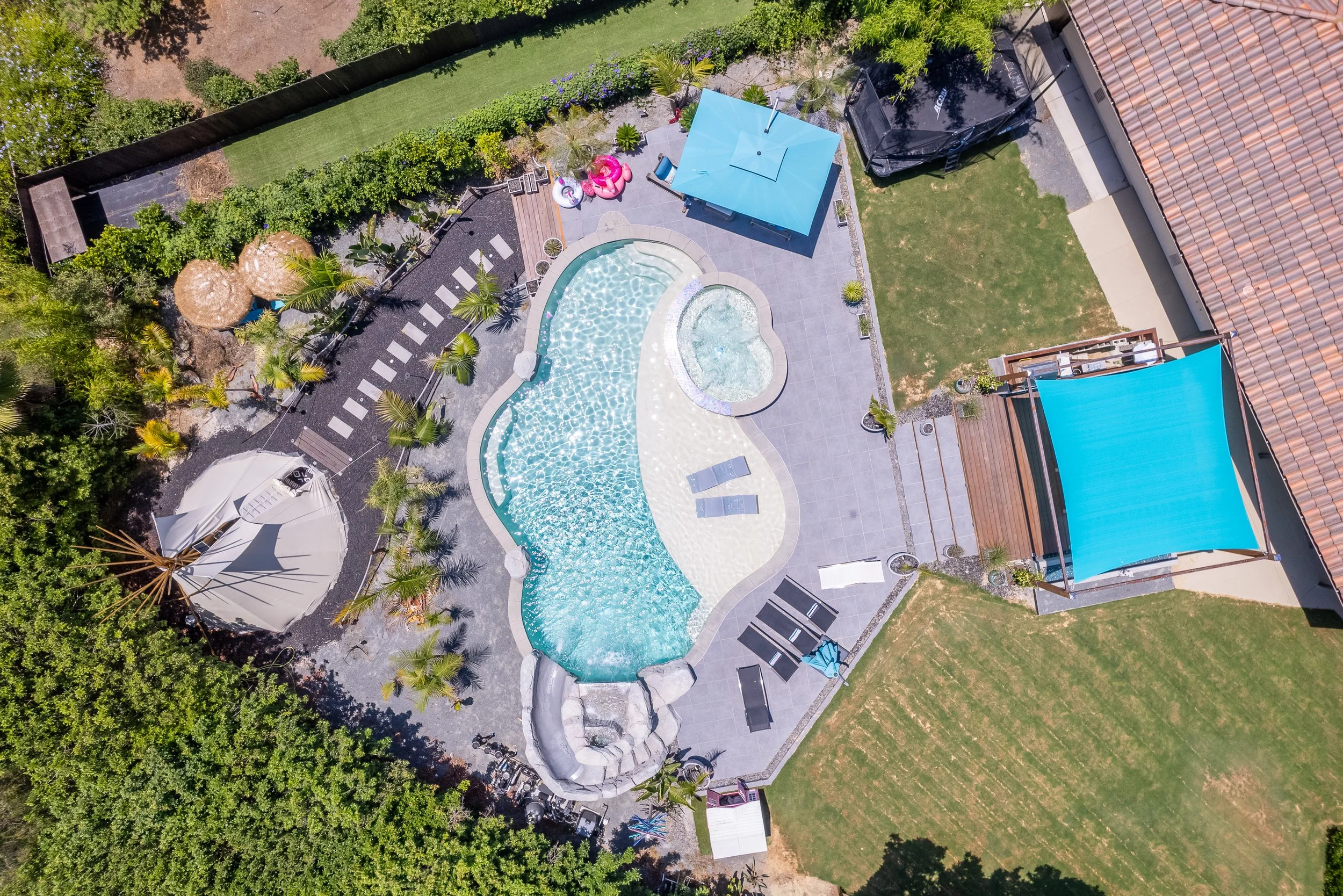 Aerial view of a backyard with a swimming pool, hot tub, shade umbrellas, lounge chairs, inflatable floaties, lush greenery, and a trampoline.