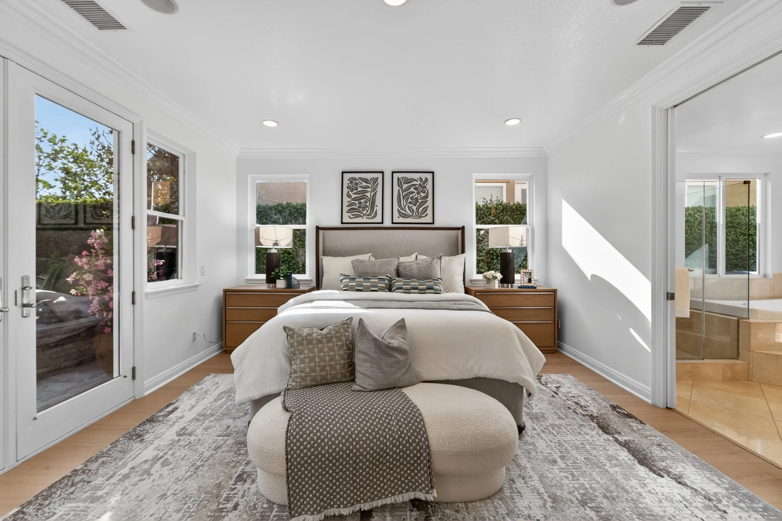 A spacious bedroom with white walls and large windows, a bed with beige bedding and decorative pillows, two bedside tables with lamps, artwork above the bed, and a patterned area rug on the wooden floor.