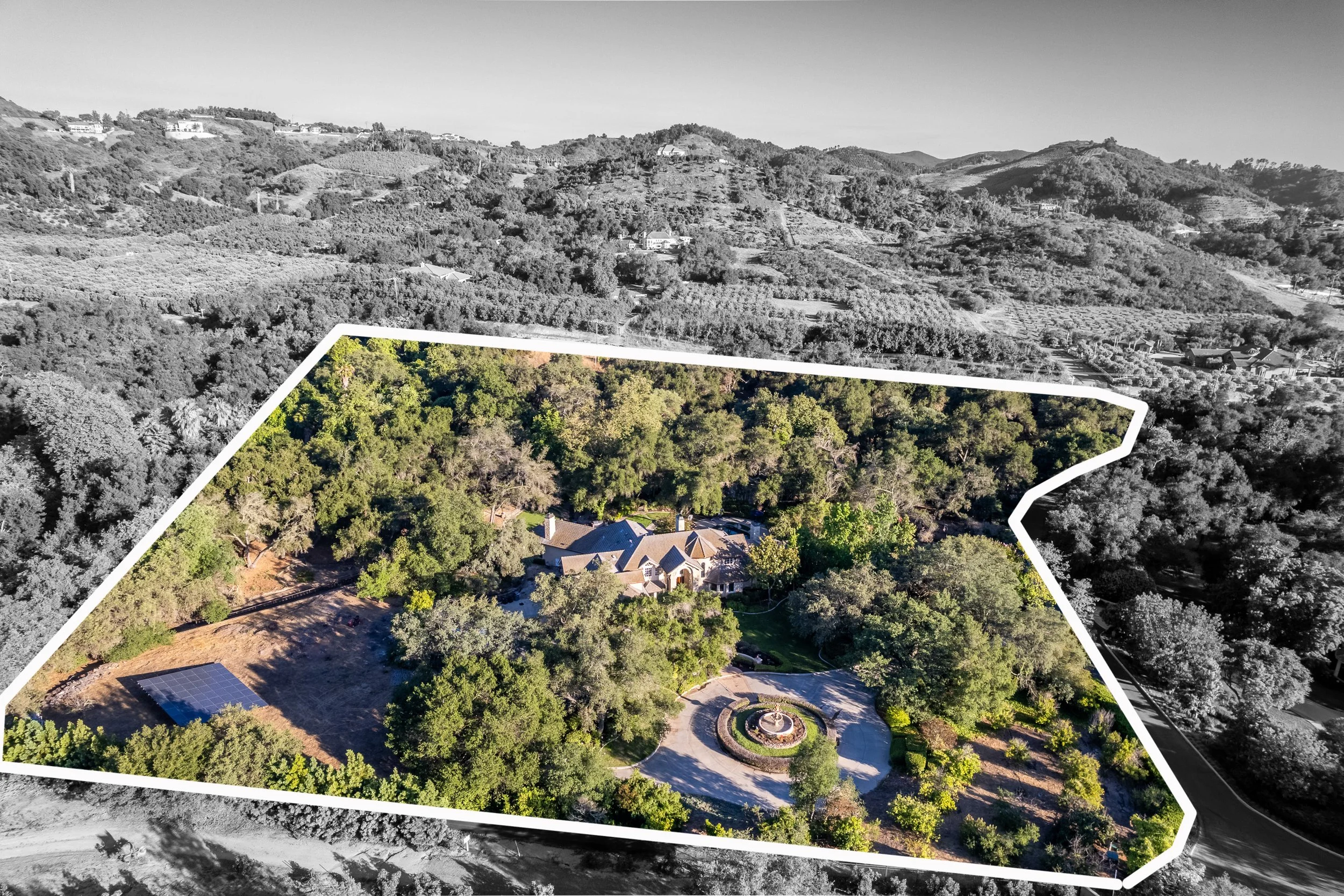 Aerial view of a residential estate with lush greenery, a circular driveway, and a fountain at the center, bordered by a white outline amidst a landscape of rolling hills and trees.