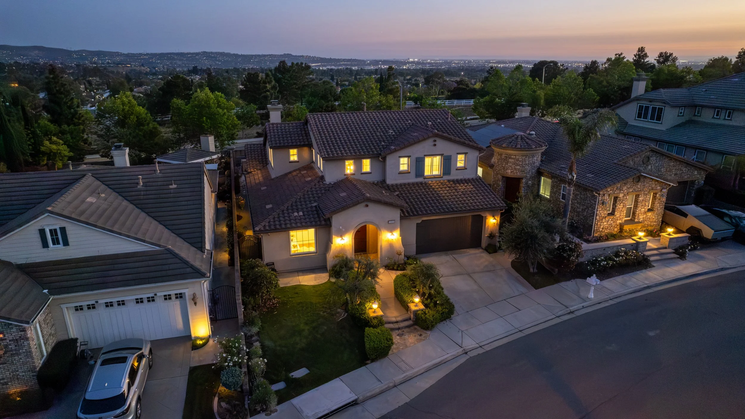 Aerial view of a suburban neighborhood at dusk with illuminated houses, tree-lined streets, and city lights in the distance.