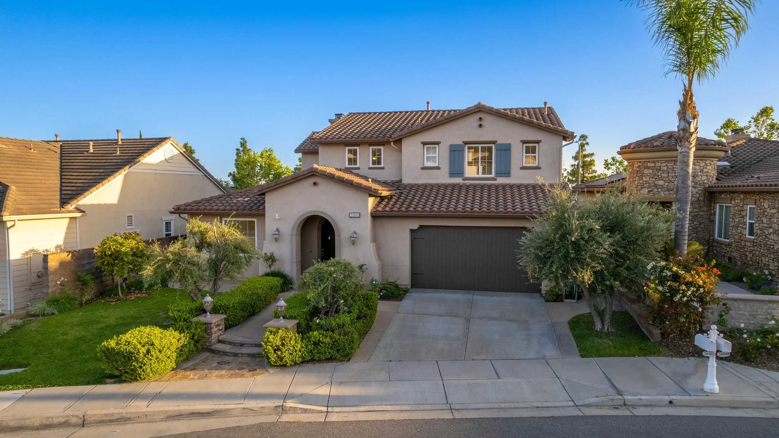 A two-story house with stucco exterior, clay tile roof, and blue shutters on the second-floor window. The front yard has neatly trimmed bushes, trees, and a sidewalk with a white mailbox. The driveway leads to a garage, and the house is surrounded by neighboring homes amidst a clear blue sky.