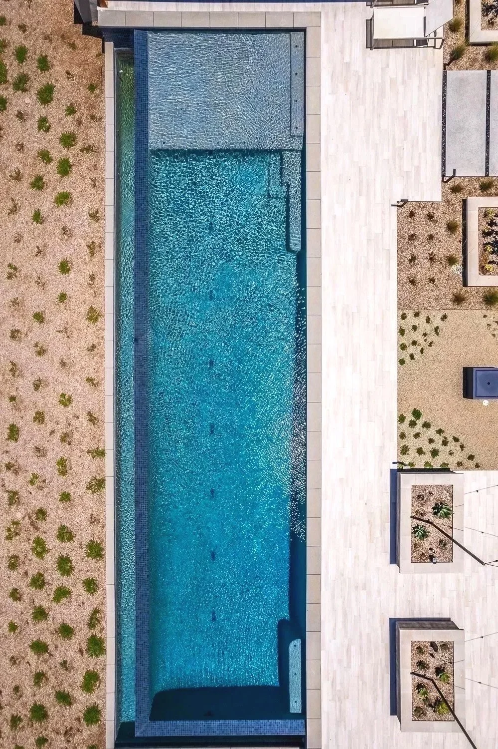 An aerial view of a rectangular swimming pool, surrounded by a white tiled deck, with landscaping featuring small bushes and desert plants on the sides.