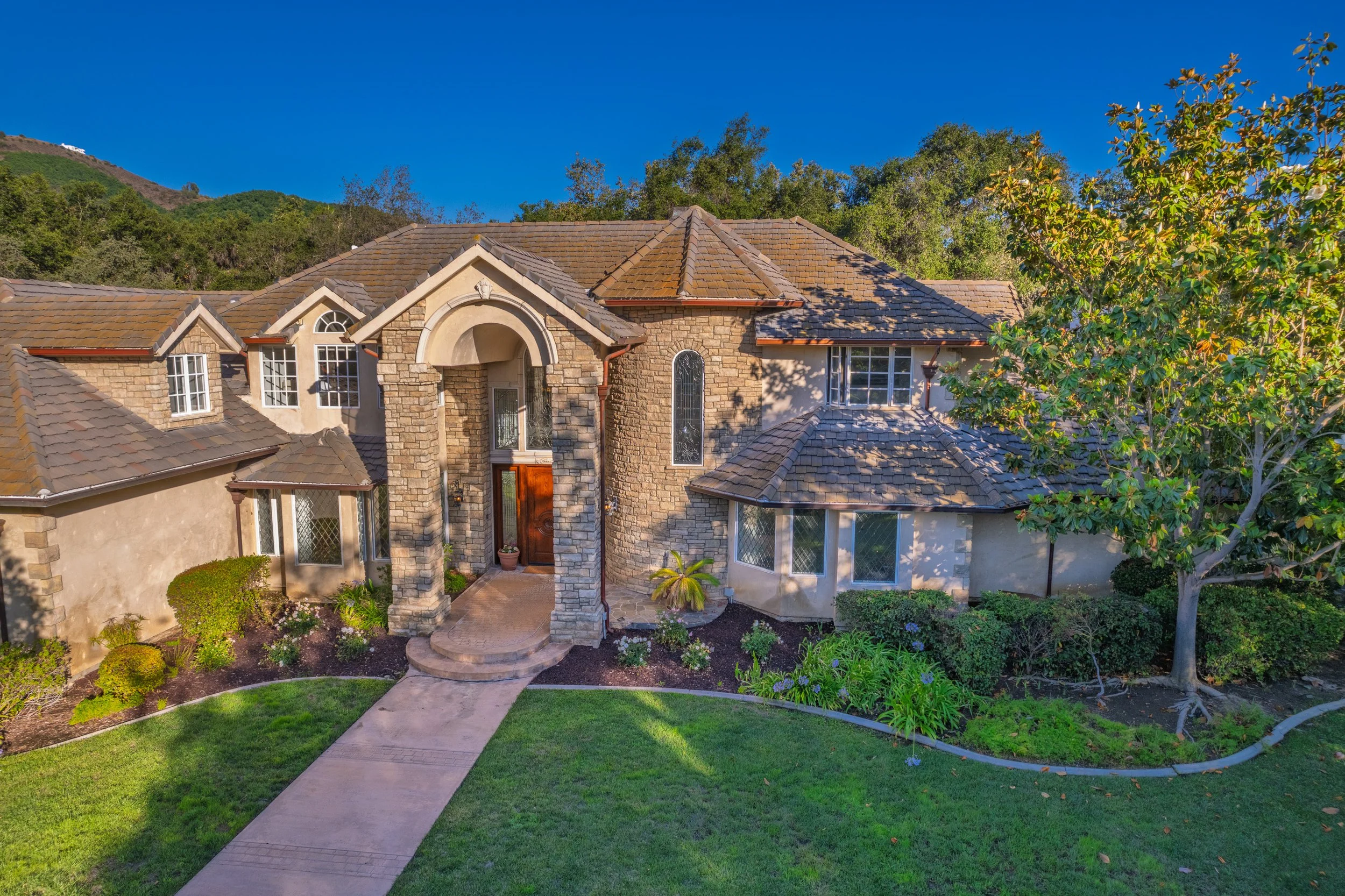 A large, two-story house with stone and stucco exterior, multiple gabled roofs with brown shingles, and a landscaped front yard with a curved pathway, green grass, shrubs, and trees.