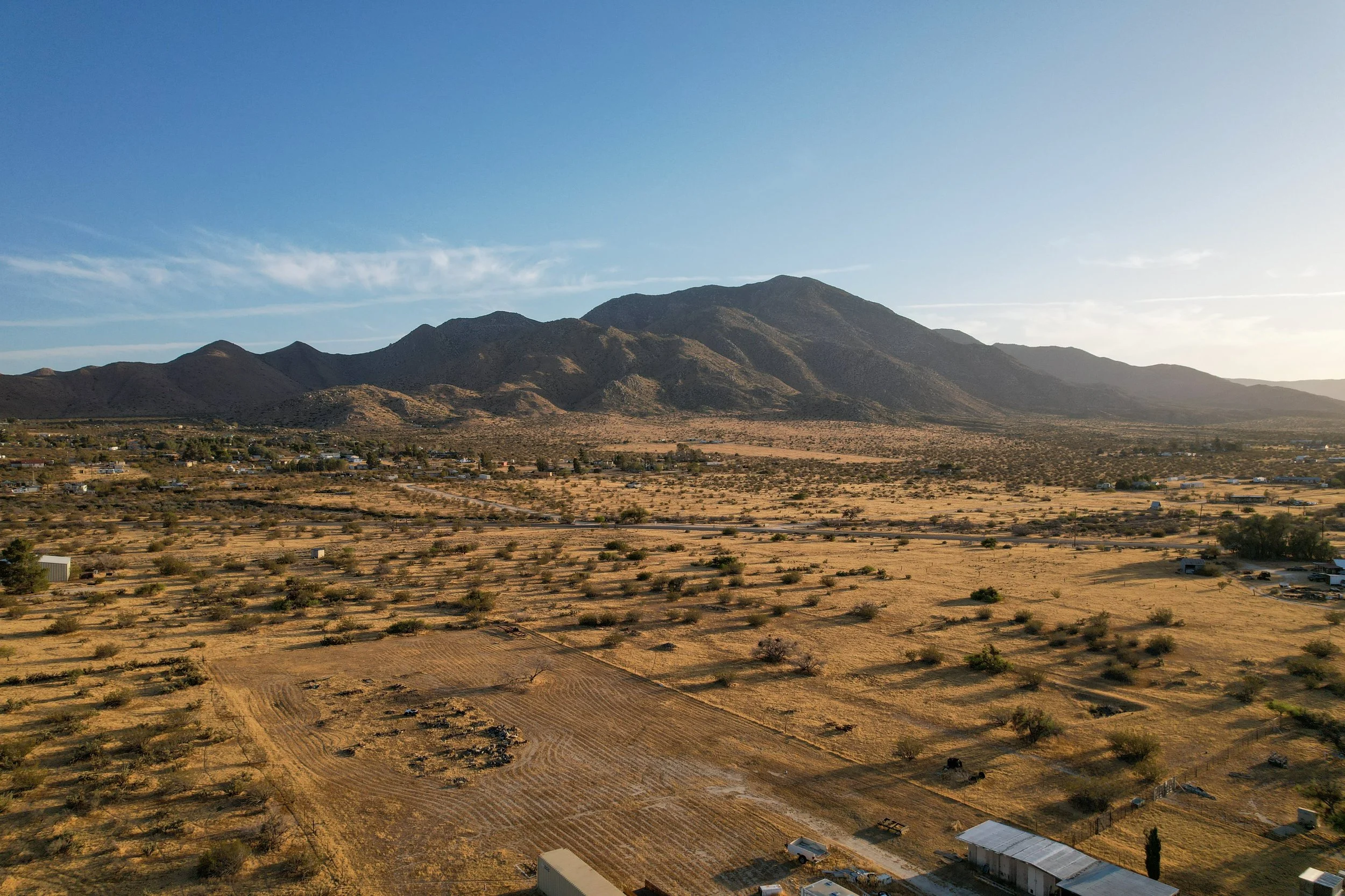A vast dry desert landscape with small scattered trees and bushes, mountain ranges in the background under a clear blue sky, and a few buildings and vehicles in the foreground.