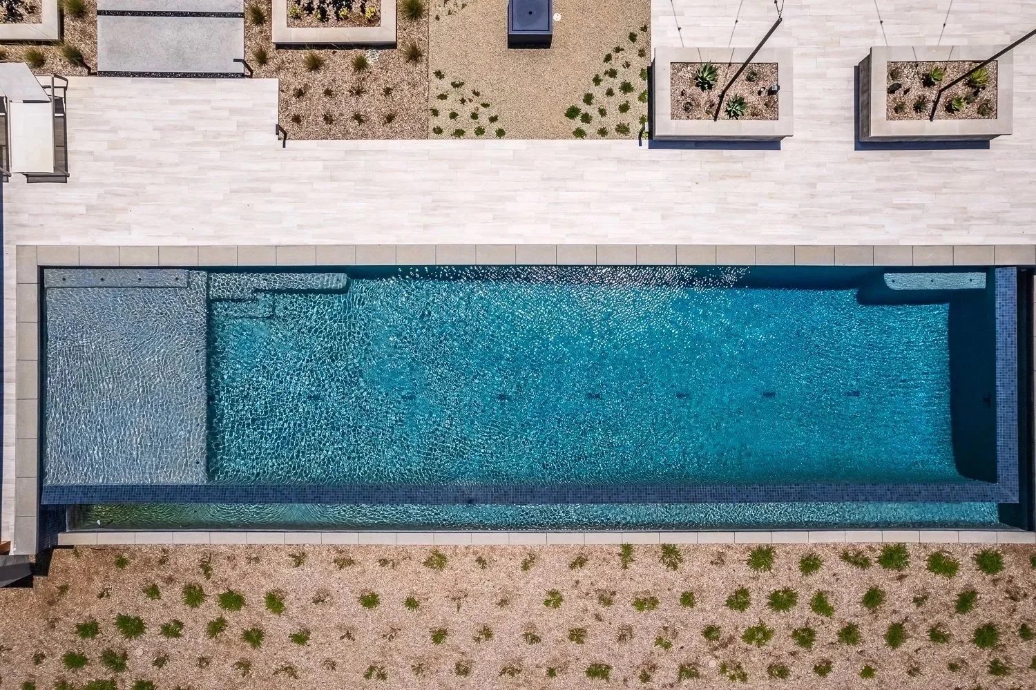 Aerial view of a modern swimming pool with a rectangular shape, water features, and a splash pad, surrounded by a beige patio and desert landscaping with small plants and bushes.