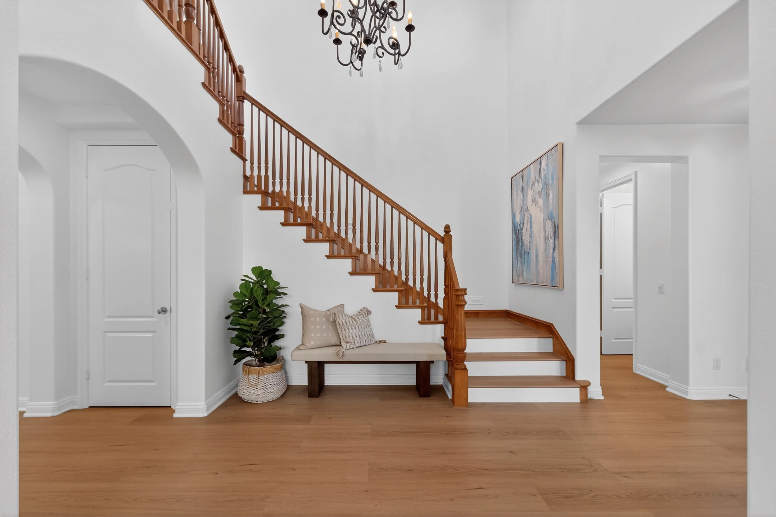 Entryway with a staircase featuring wooden handrails and spindles, a black chandelier hanging from the ceiling, a framed abstract painting on the wall, a bench with cushions, a potted plant, and light-colored walls with hardwood flooring.
