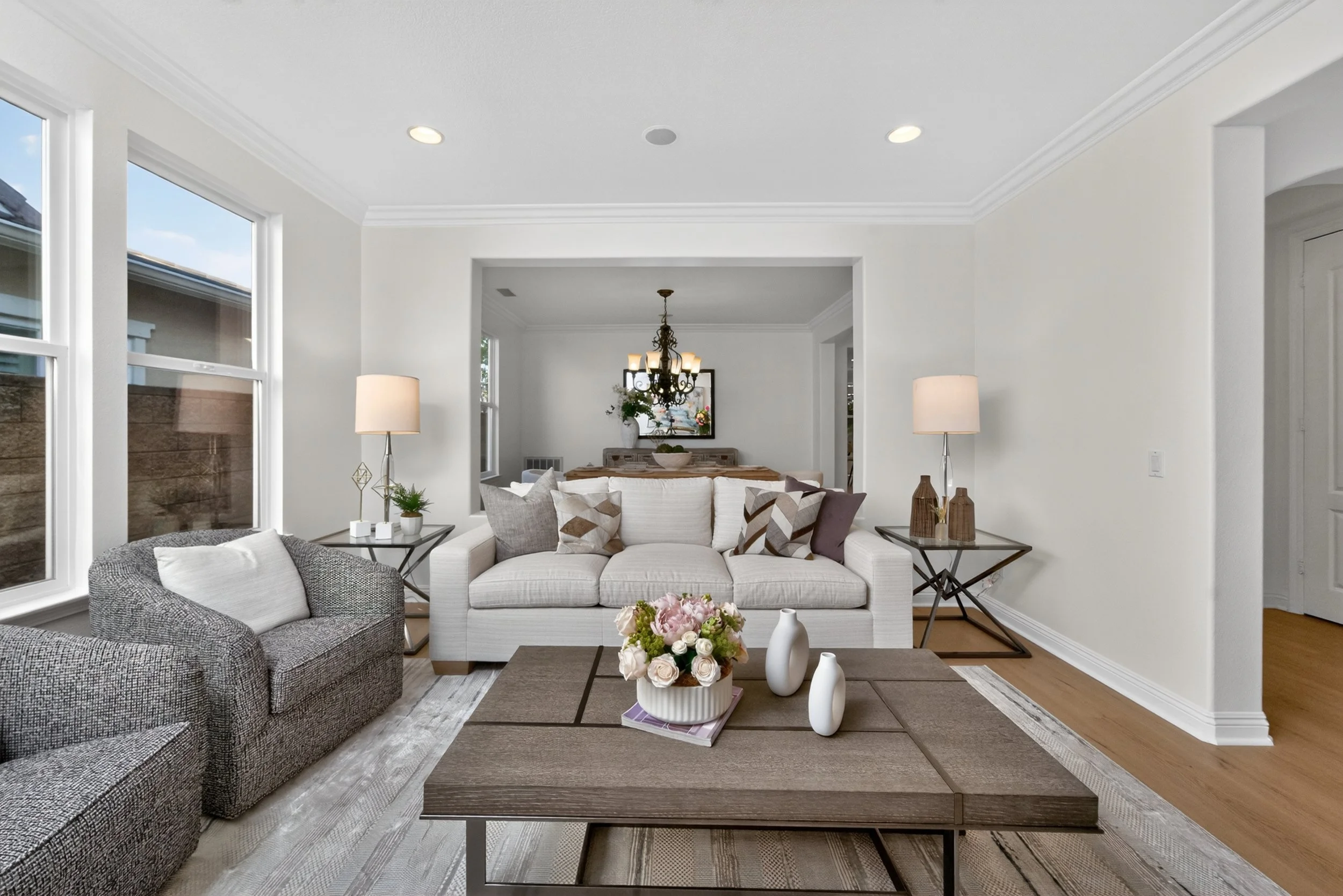 Living room with two sofas, a coffee table with a flower arrangement, and a large window with sunlight. In the background, there is a dining area with a chandelier and a mirror.