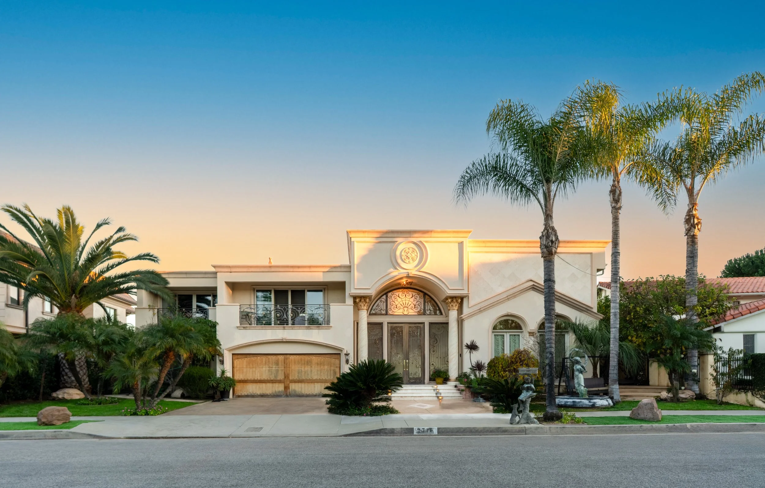 Luxury two-story house with palm trees, a gated entrance, statue decorations, and a driveway, set against a sunset sky.