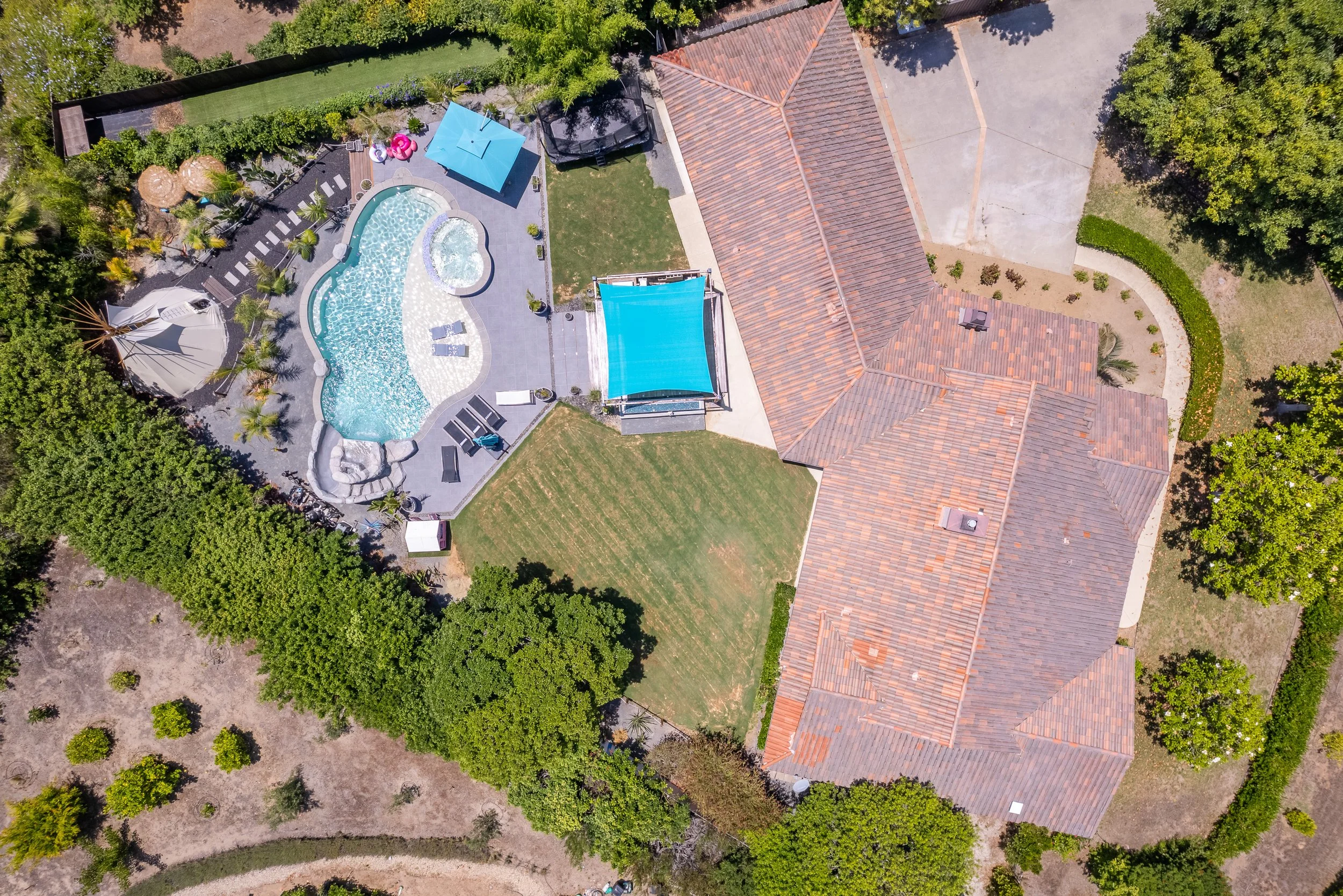 An aerial view of a backyard featuring a swimming pool with a hot tub, surrounded by a paved patio with lounge chairs, a blue shade sail, and some umbrellas. The yard has green grass, trees, plants, and a garden area enclosed by a fence. Adjacent to 