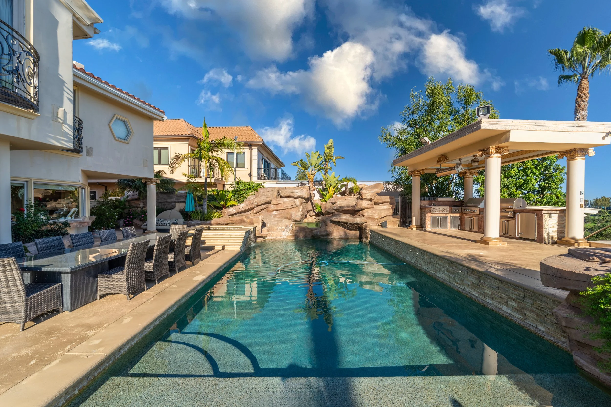 Luxury backyard with swimming pool, outdoor dining area, and poolside bar with columns, surrounded by palm and other trees under a partly cloudy blue sky.