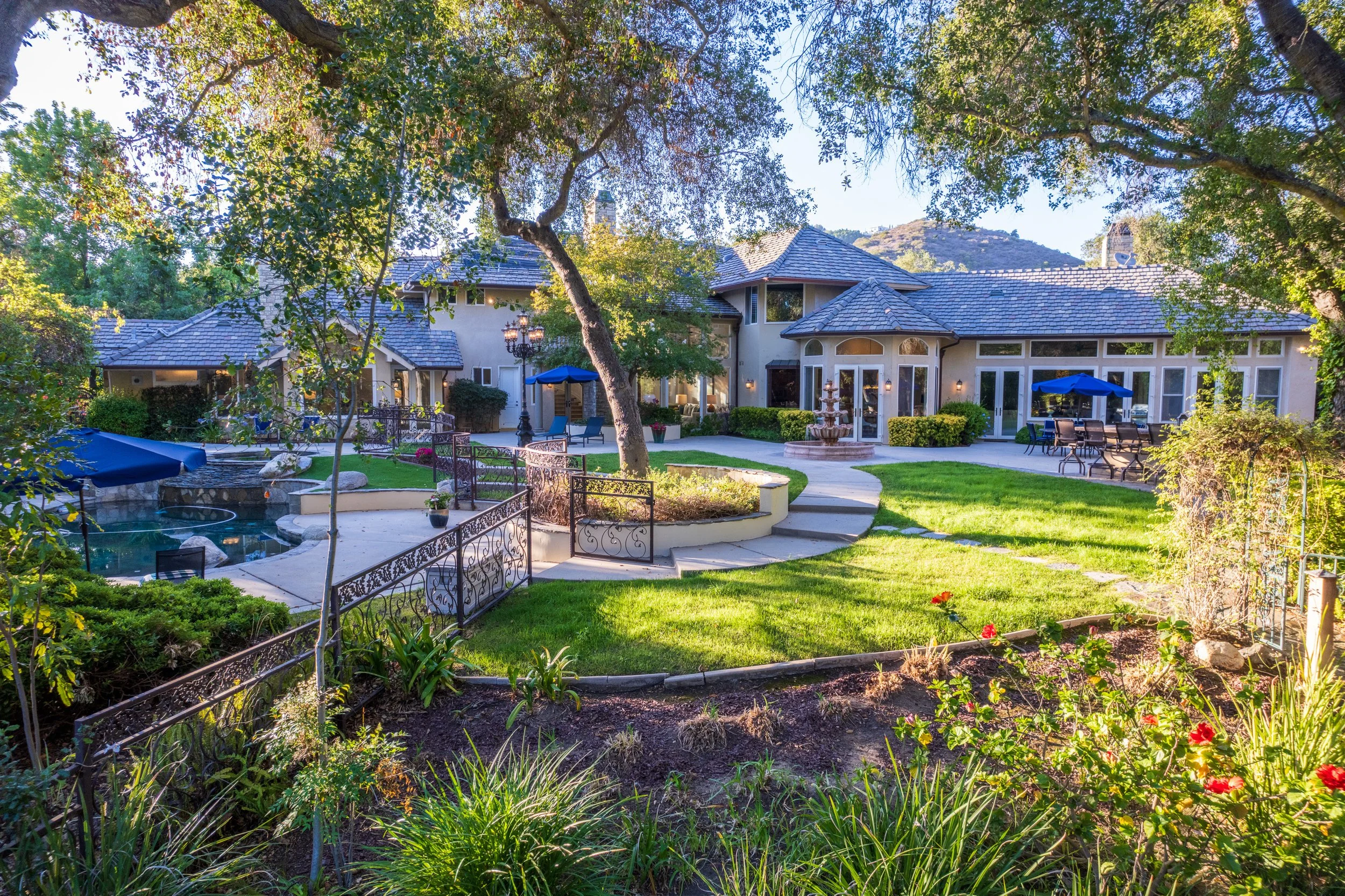 A spacious backyard featuring a house with a tiled roof, surrounded by greenery and trees. There is a paved patio area with outdoor furniture, umbrellas, and a fountain, with mountain hills visible in the background.