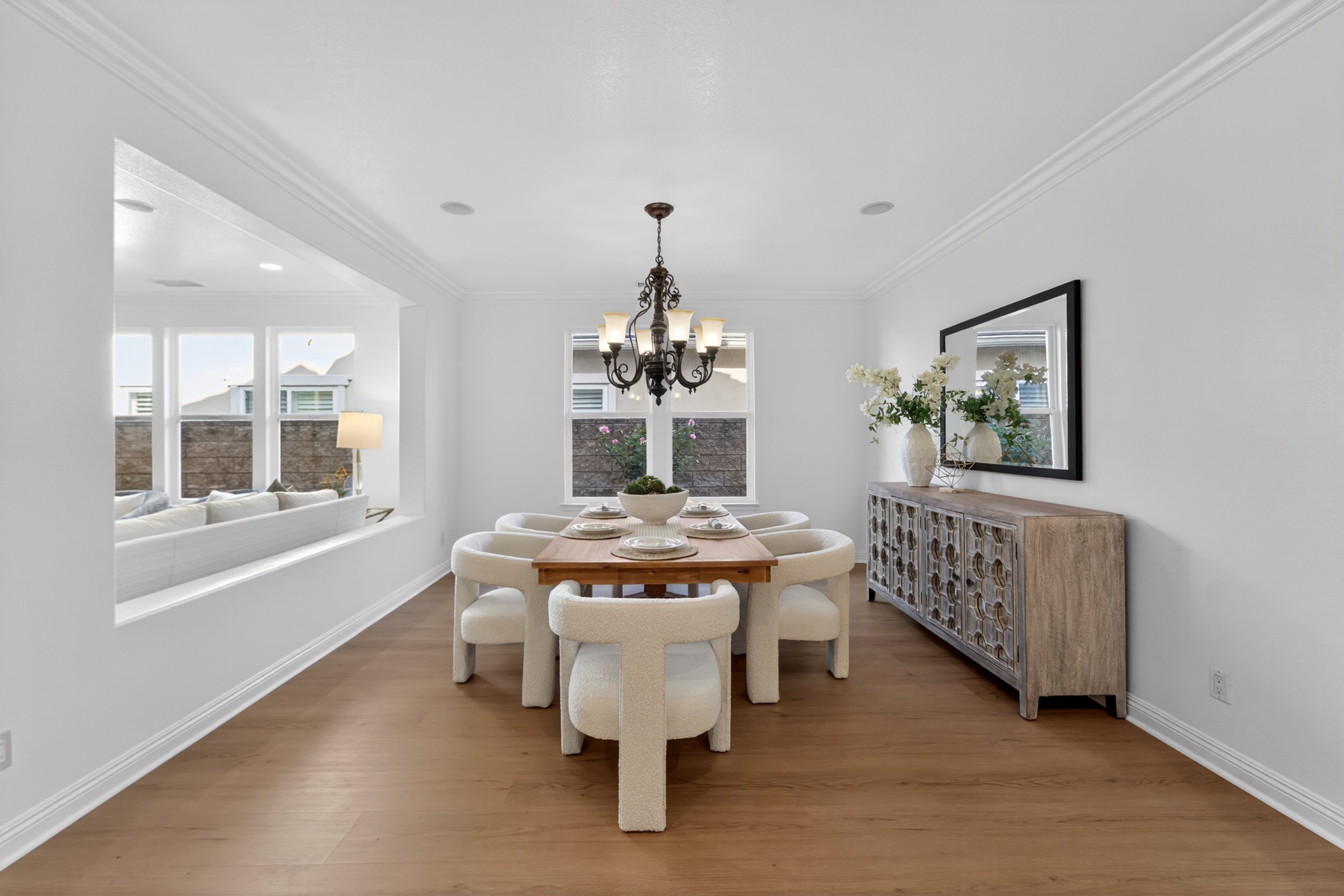 A dining room with a wooden table set with plates and six cream-colored chairs. A black chandelier hangs above the table. There is a sideboard with decorative vases and flowers, and a large mirror on the wall. Windows in the background let in natural