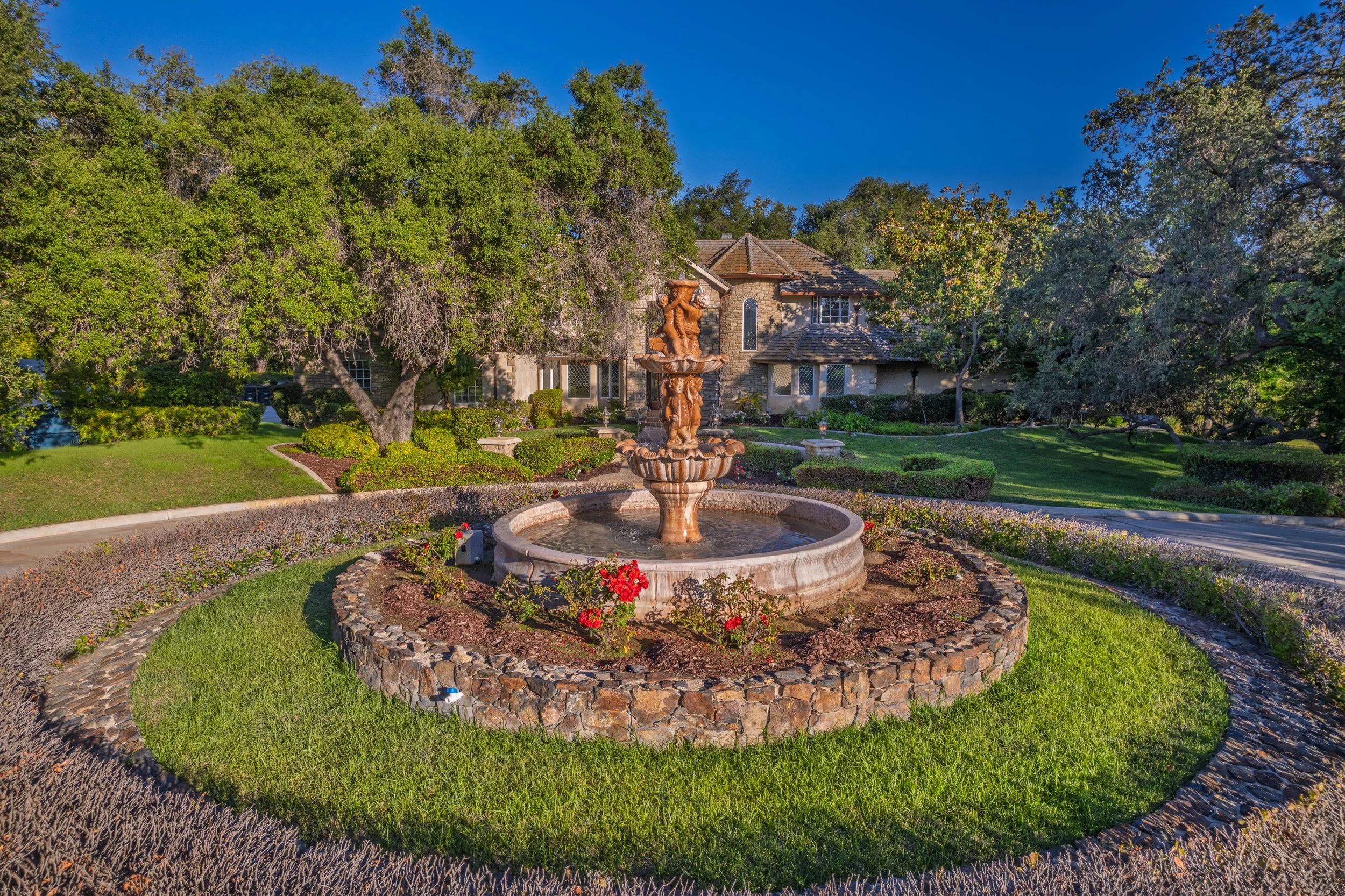 A garden with a stone fountain in the center, surrounded by colorful flowers and trimmed bushes, with a large house and mature trees in the background under a blue sky.
