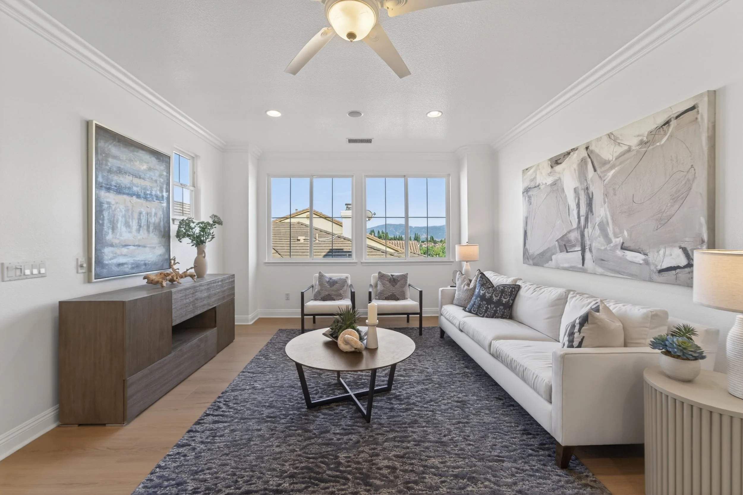 Living room with white walls, large window showing a view of rooftops and mountains, ceiling fan, abstract paintings, a white sofa with patterned pillows, two armchairs, a round coffee table with decor, a side table with a lamp, and a dark patterned 