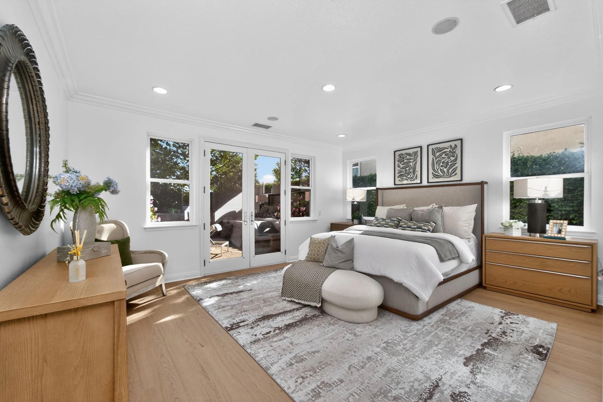 A bright living room with a large white bed, wooden side tables, and decorative black and white artwork. Large windows and a glass door letting in natural light, with visible outdoor greenery and patio furniture.