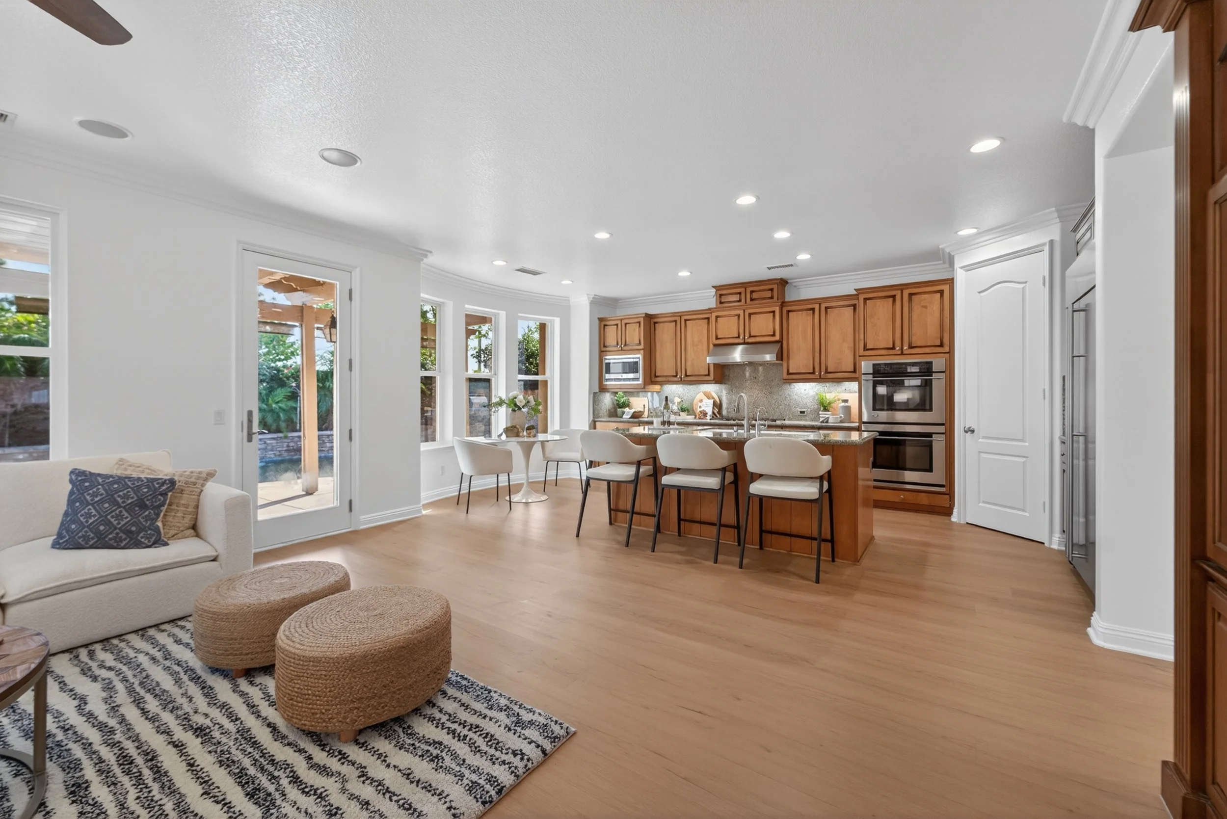 Interior view of a modern kitchen and living room with wooden cabinets, a dining table with chairs, bar stools, and a patterned rug. The space has white walls, large windows, and hardwood floors.