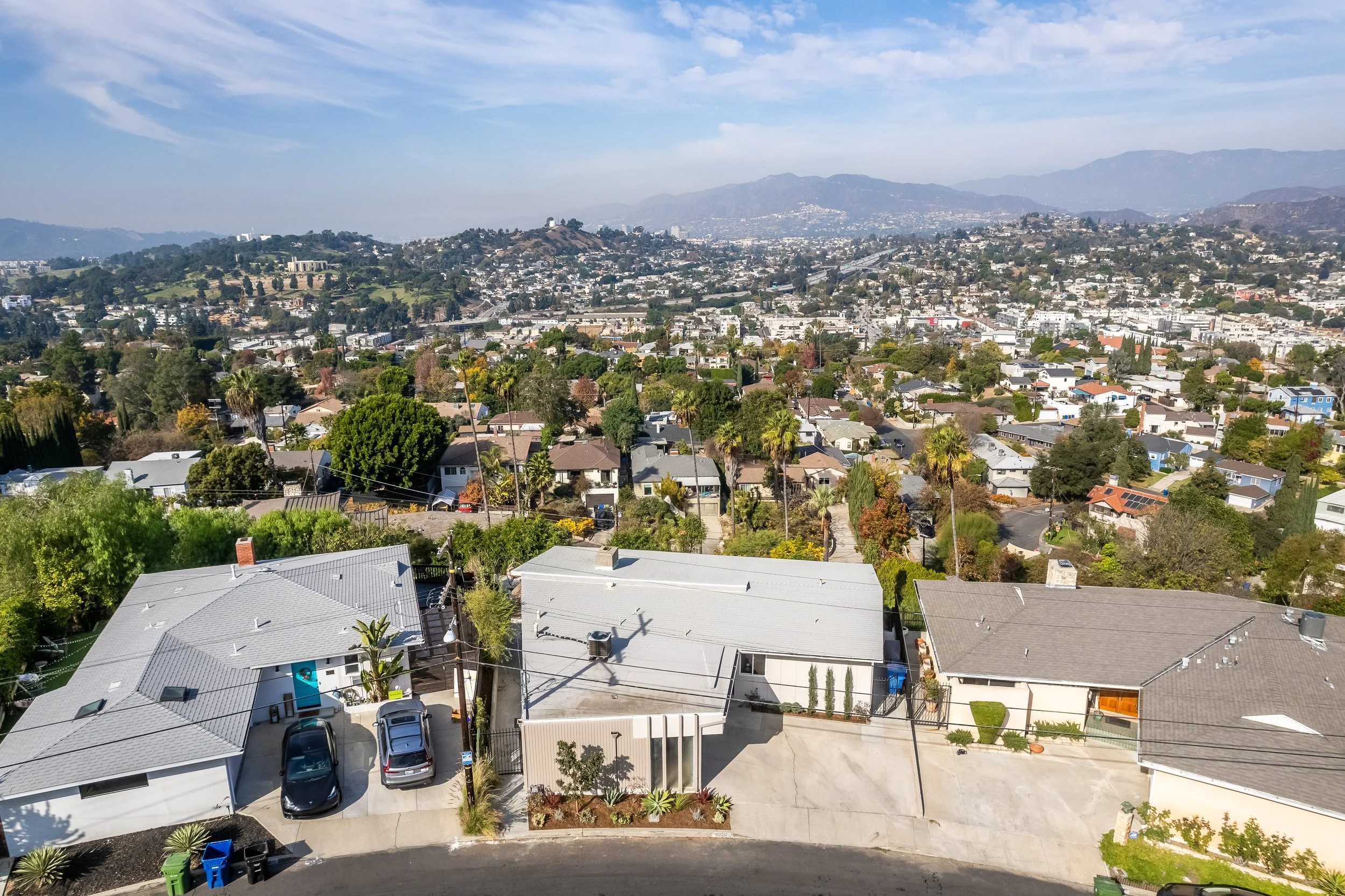 Aerial view of a residential neighborhood with houses, driveways, and trees, overlooking a cityscape and distant mountains under a partly cloudy sky.